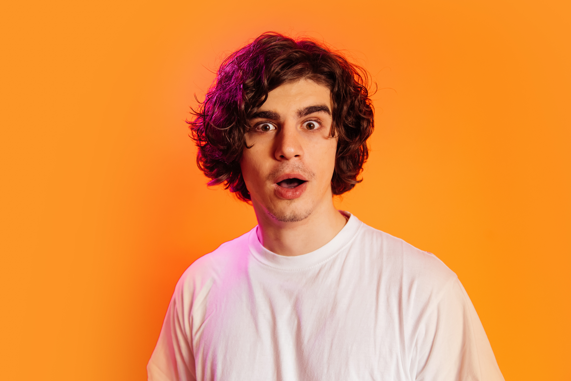 A young man with curly hair and a surprised expression stands against an orange background, wearing a white t-shirt.