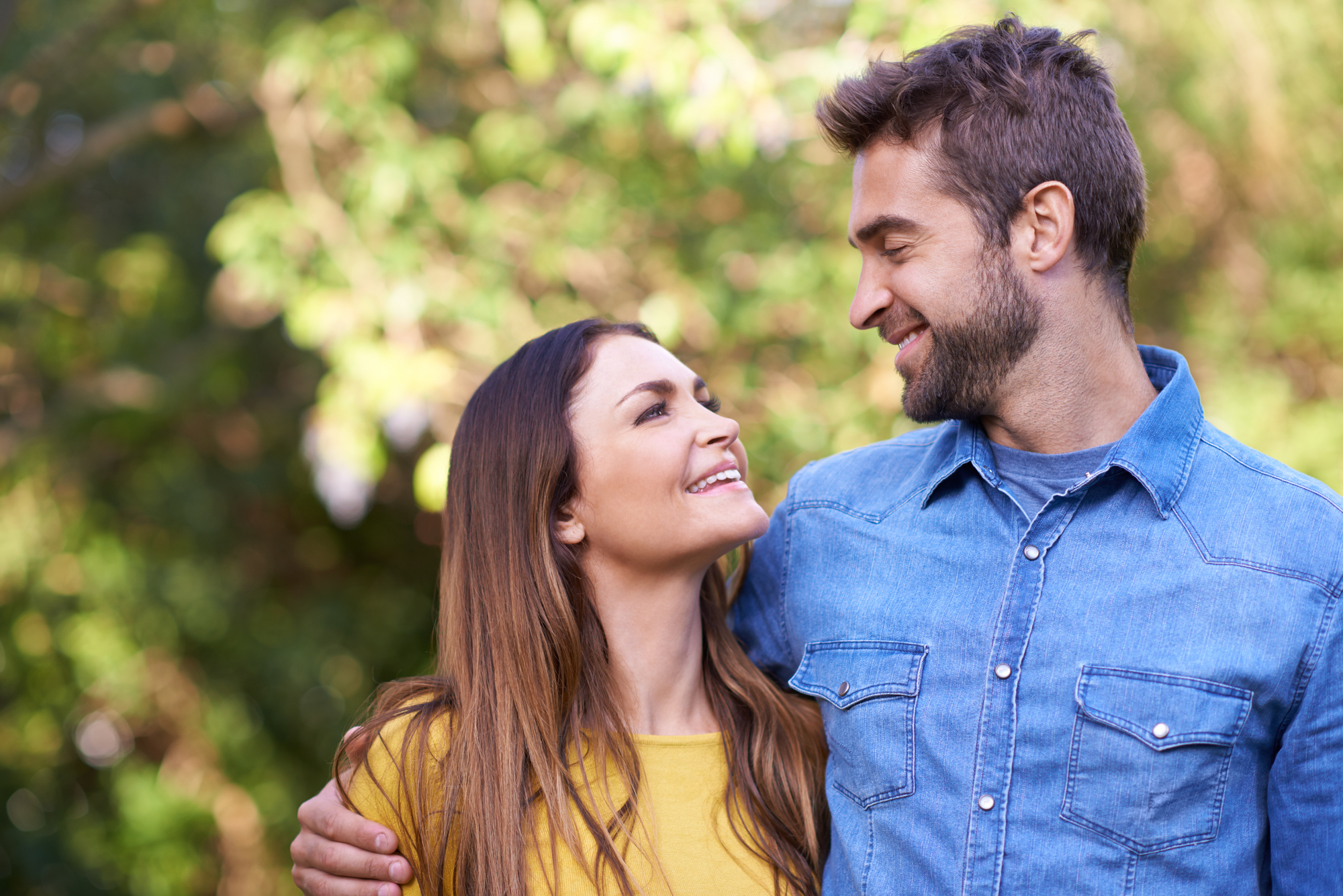 A couple stands outside, smiling at each other with trees in the background. The woman wears a yellow top, and the man wears a denim shirt, with his arm around her shoulders. Both appear happy and relaxed.