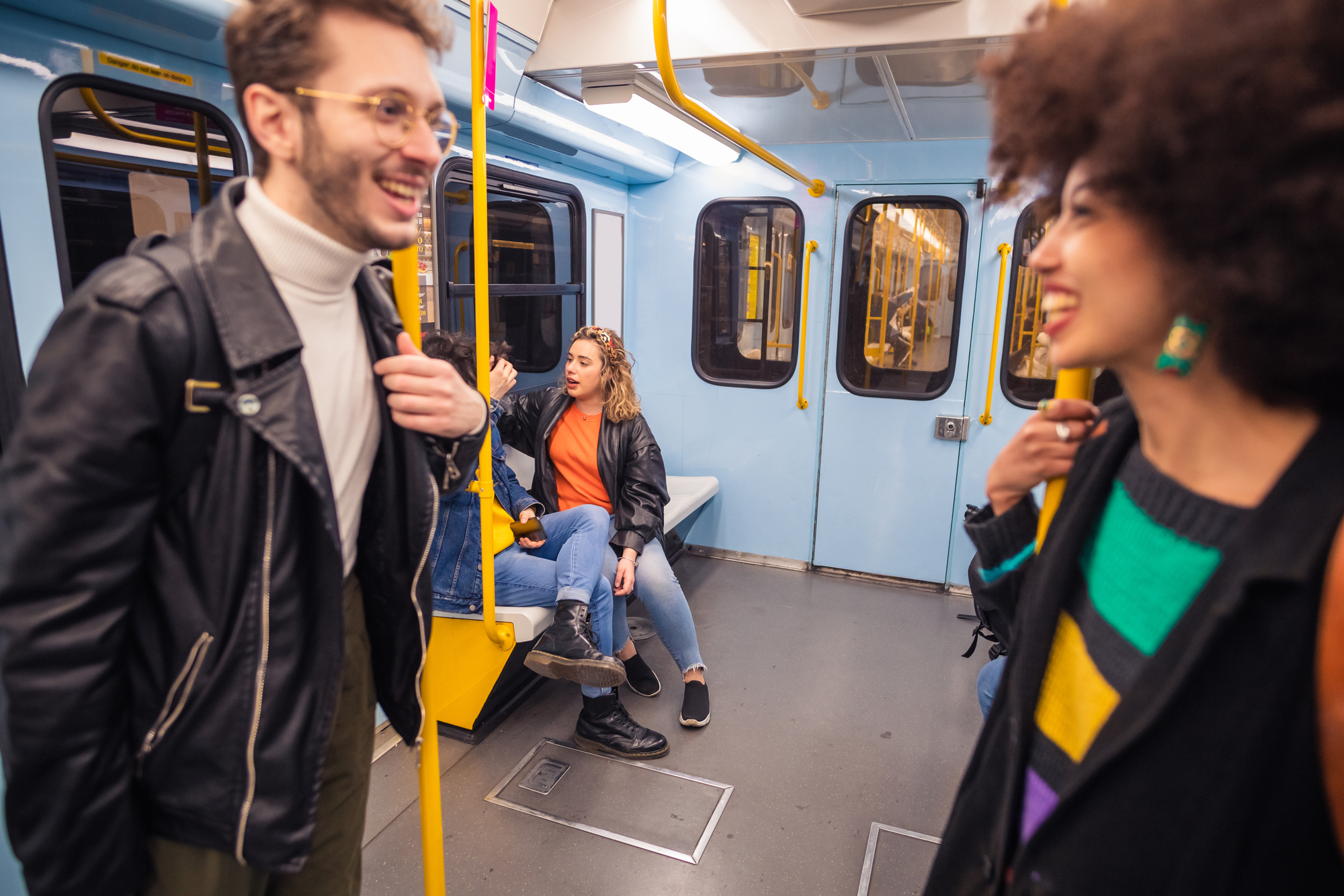 A group of young adults in a subway car. Two people are standing and smiling in the foreground, holding onto poles. Two others are seated, engaged in conversation. The subway interior is light blue with yellow accents.