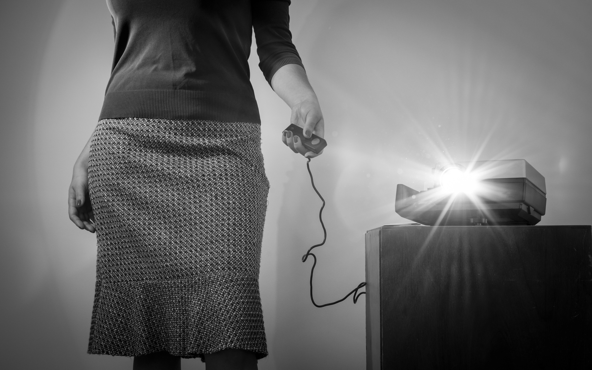 A person stands beside a slide projector, holding a remote control, with the projector casting a bright light. The image is in black and white, focusing on the lower half of the person's body and the equipment.