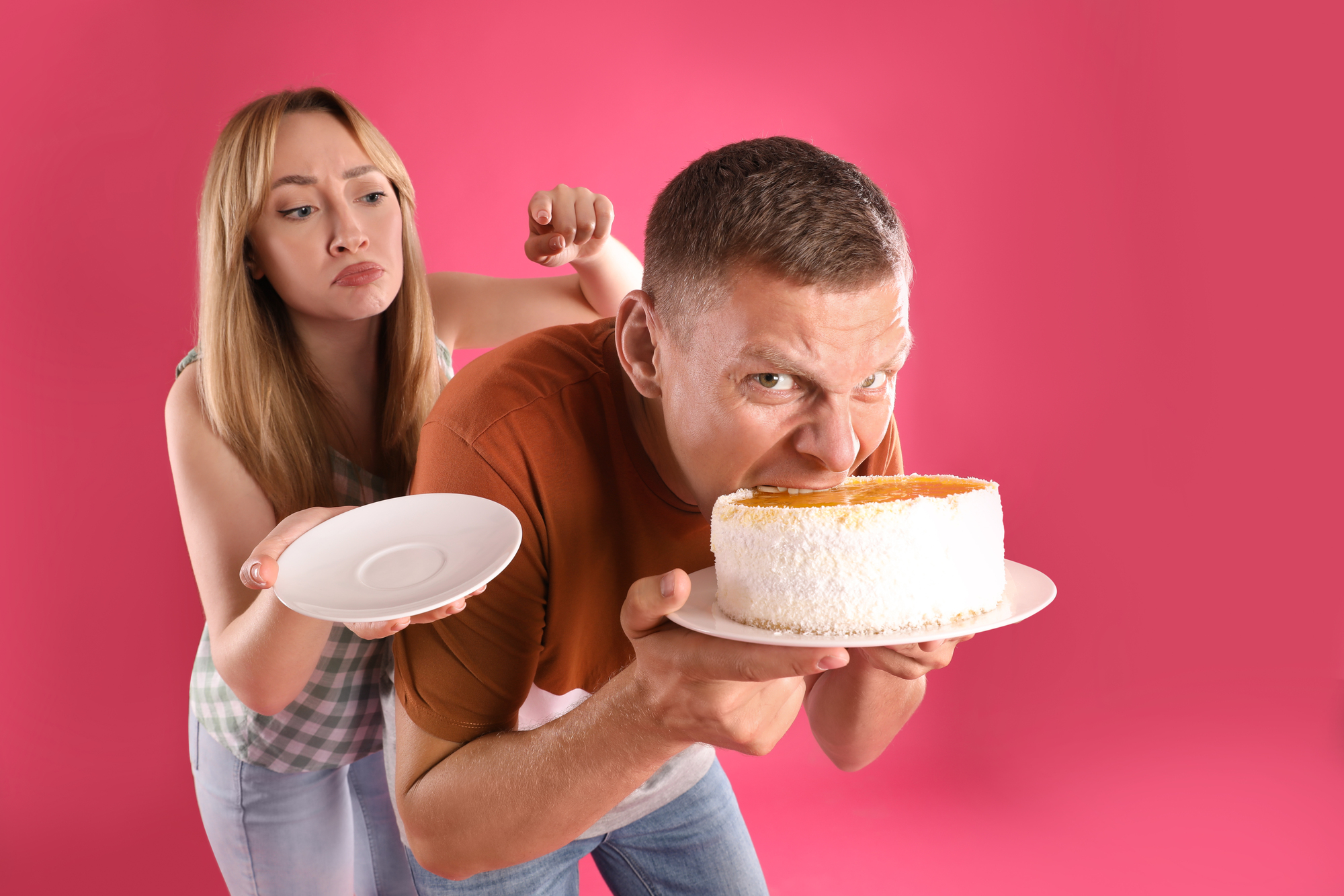 A man humorously eats a cake directly with his mouth while holding the plate. A woman beside him looks at the empty plate she's holding, pouting in mock disappointment. The background is bright pink.