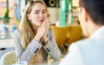 A woman with long blonde hair, wearing a light gray blazer, sits at a table with her hands clasped, appearing thoughtful. She faces a person with short dark hair in a blurred foreground. A cup is on the table in a bright, modern setting.