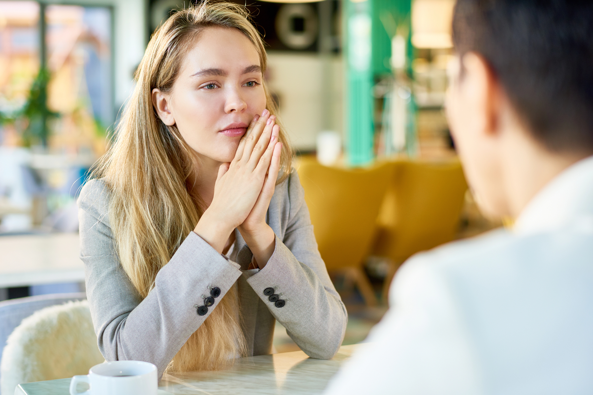 A woman with long blonde hair, wearing a light gray blazer, sits at a table with her hands clasped, appearing thoughtful. She faces a person with short dark hair in a blurred foreground. A cup is on the table in a bright, modern setting.