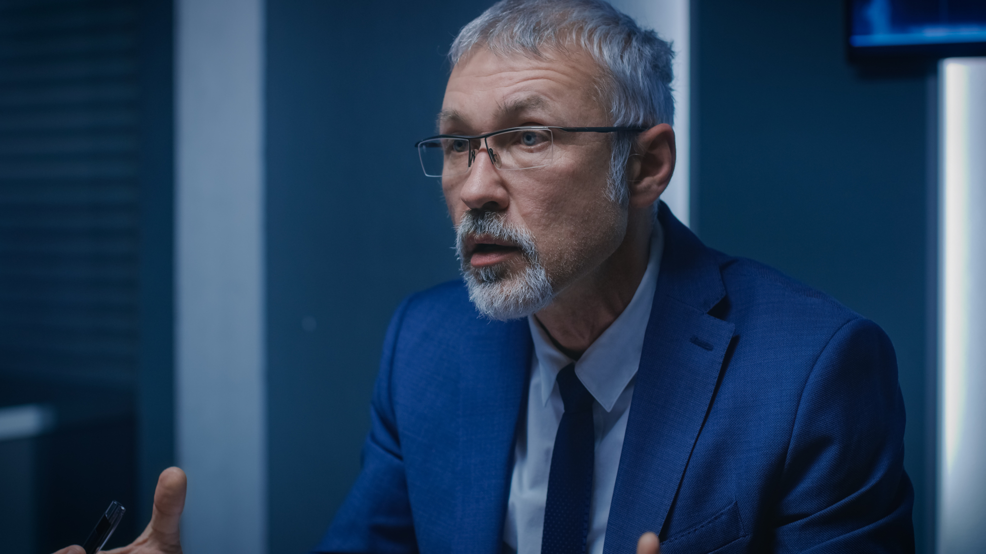 An older man with gray hair and a beard is wearing glasses and a blue suit. He appears to be speaking or engaged in a conversation against a dark, blurred background.