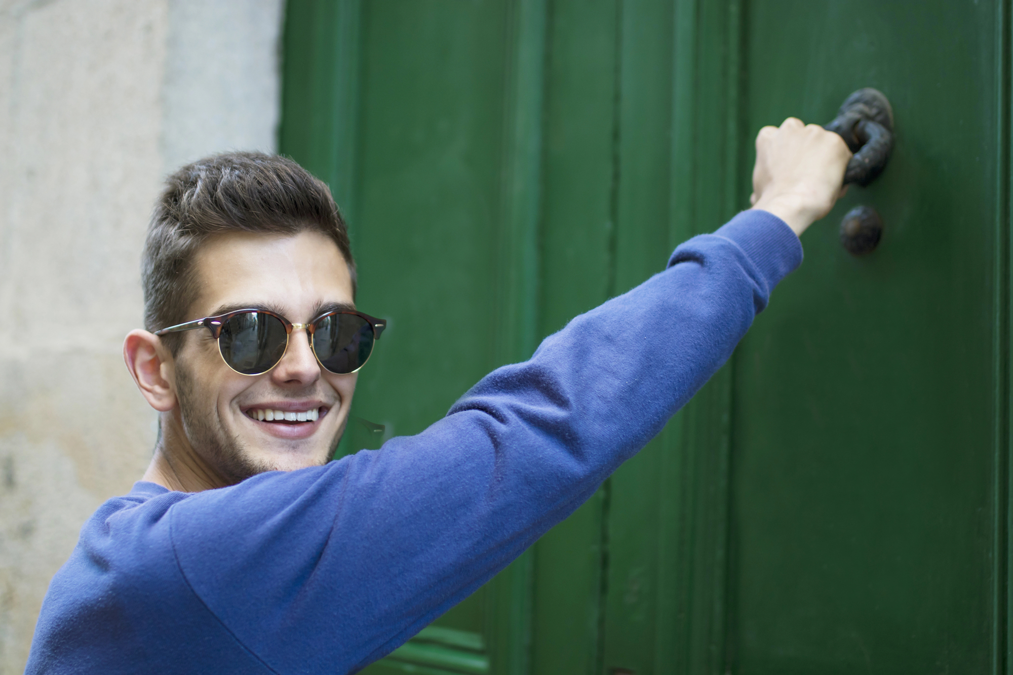 A person wearing sunglasses and a blue sweater is smiling while knocking on a green door.