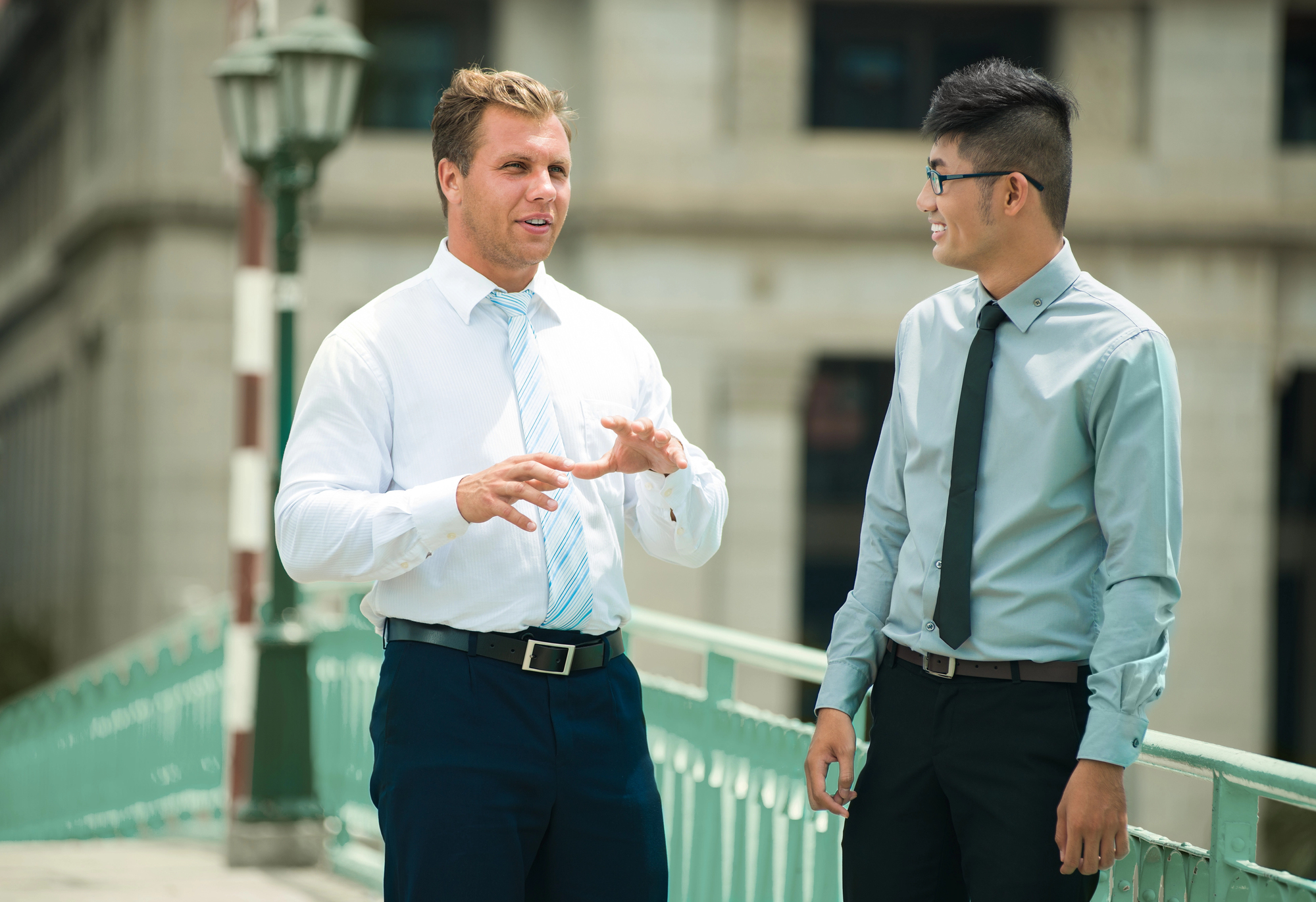 Two men in professional attire are having a conversation while standing on a bridge with a vintage-style lamp post. One man gestures with his hands, and they both appear engaged and smiling. The background shows a blurred building.