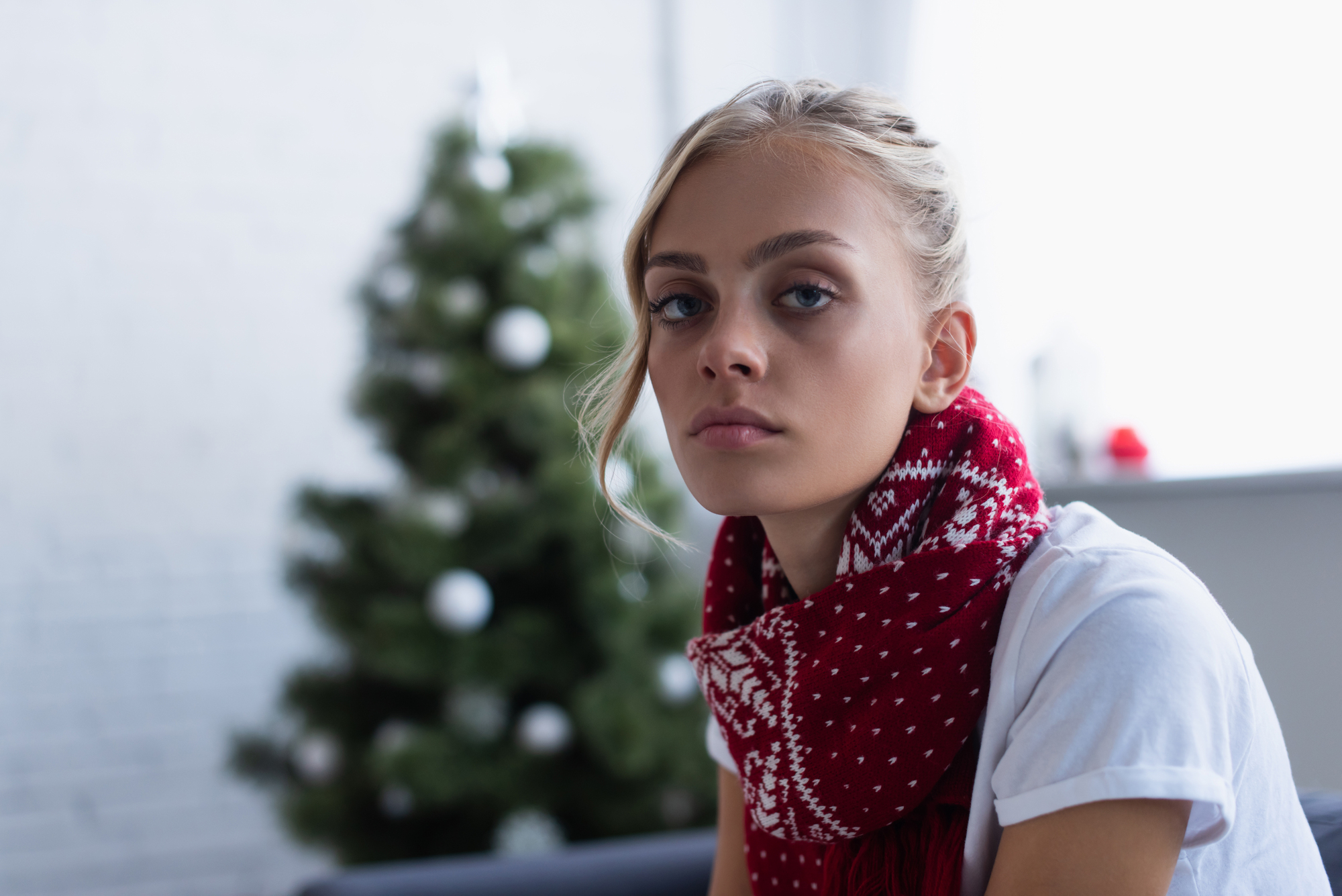 A person with long blonde hair wears a red and white scarf and a white shirt, sitting indoors. In the background, there is a blurred Christmas tree decorated with white ornaments. The scene has a calm and festive atmosphere.