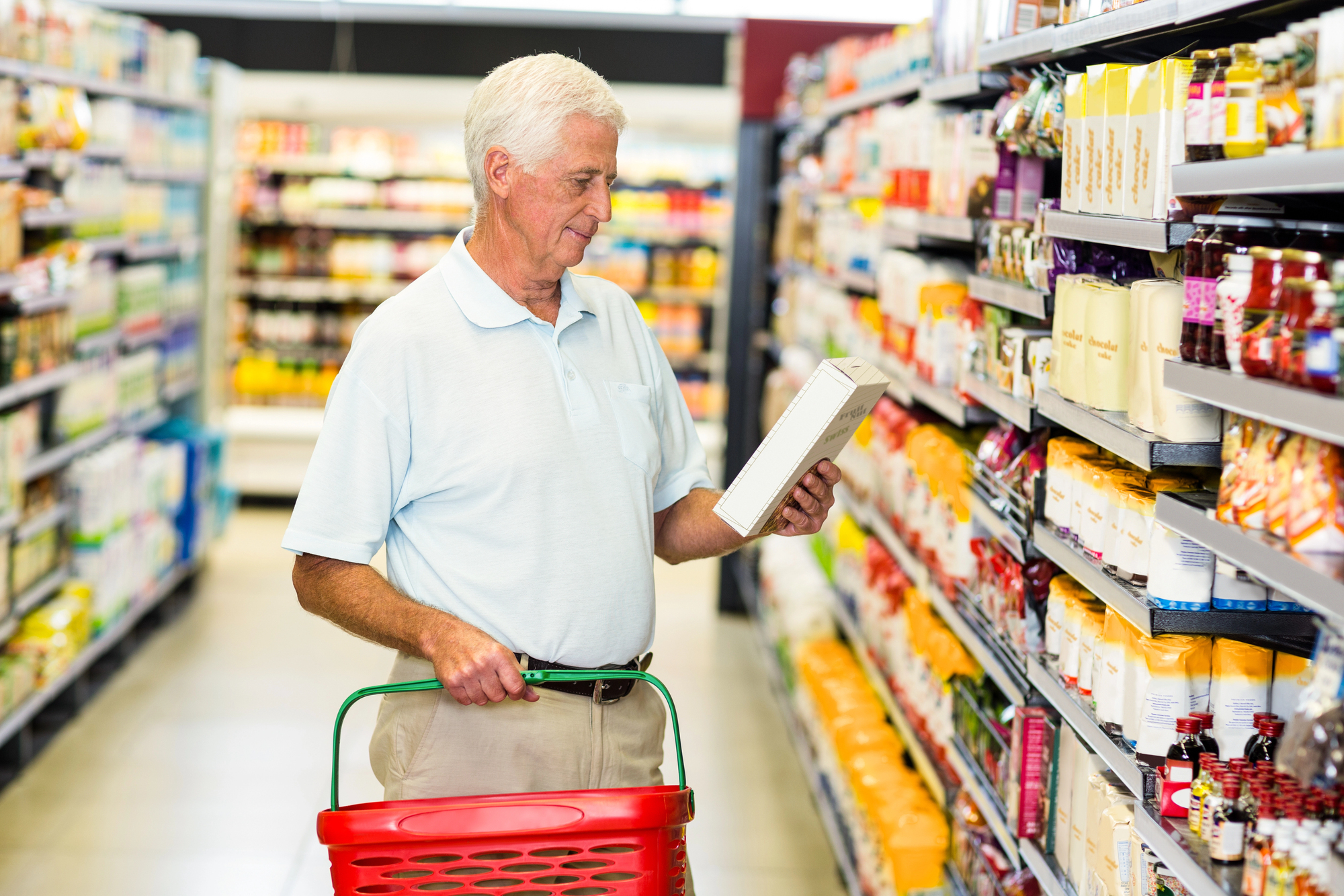 Elderly man with short white hair, wearing a light blue polo shirt, holds a red shopping basket and examines a product in a grocery store aisle. Shelves stocked with various goods line both sides.