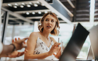 A woman in a sleeveless top sits at a table with a laptop in front of her, holding a pen. She appears engaged in a conversation with someone off-camera. The setting seems to be a bright, modern indoor space.