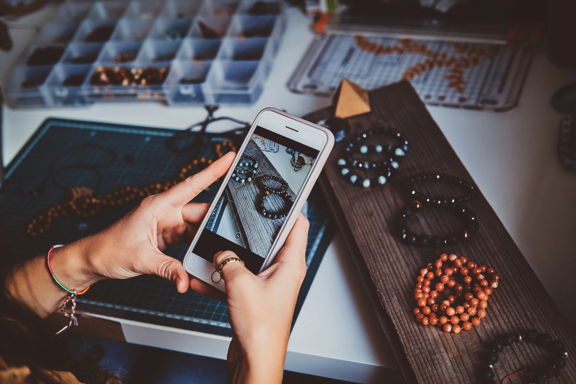 A person holds a smartphone taking a photo of handmade beaded jewelry laid out on a table. The workspace includes trays with assorted beads and crafting tools.