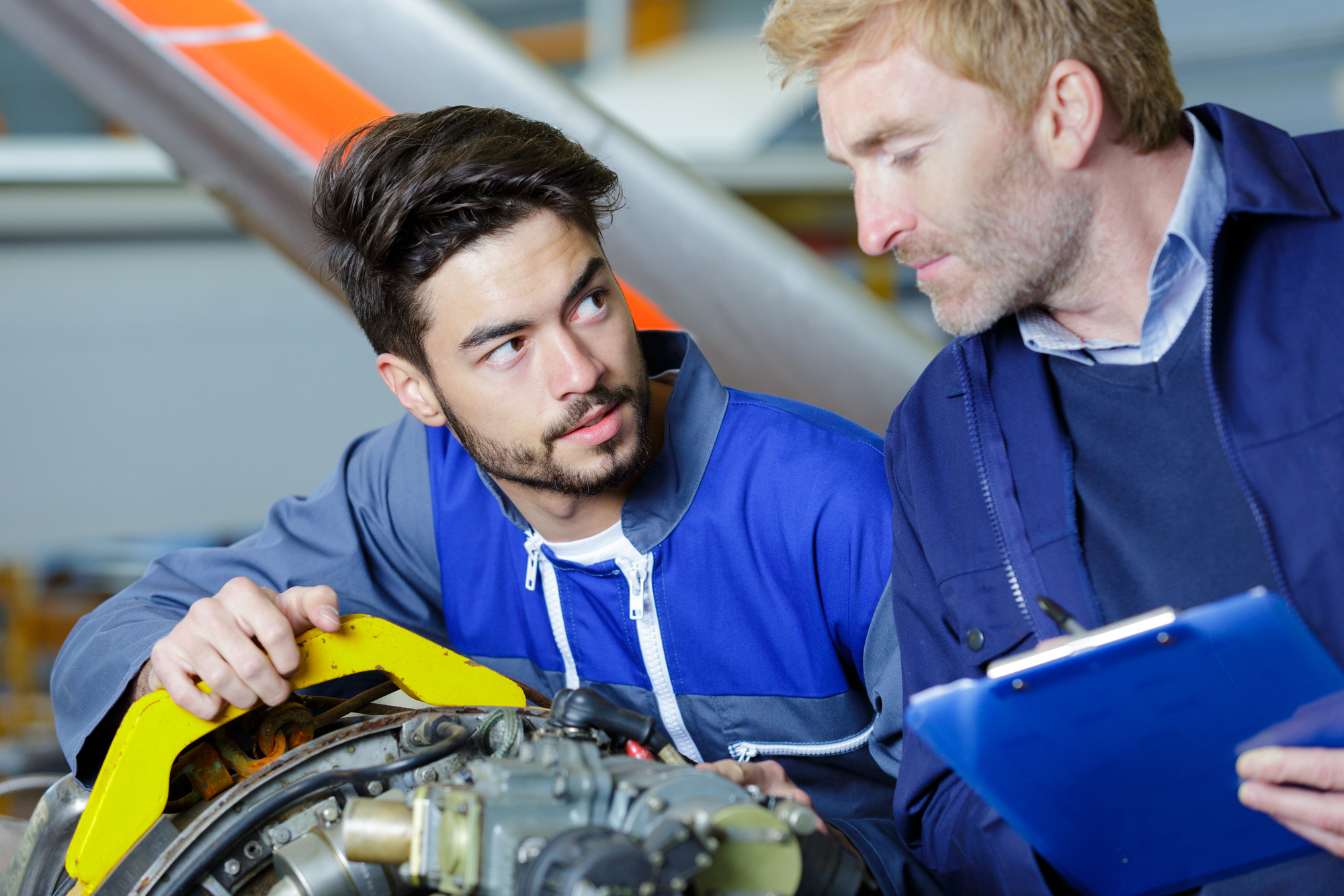 Two technicians in blue uniforms are inspecting an aircraft engine. One holds a clipboard while the other examines the engine closely. They appear to be discussing the machinery in a workshop setting.