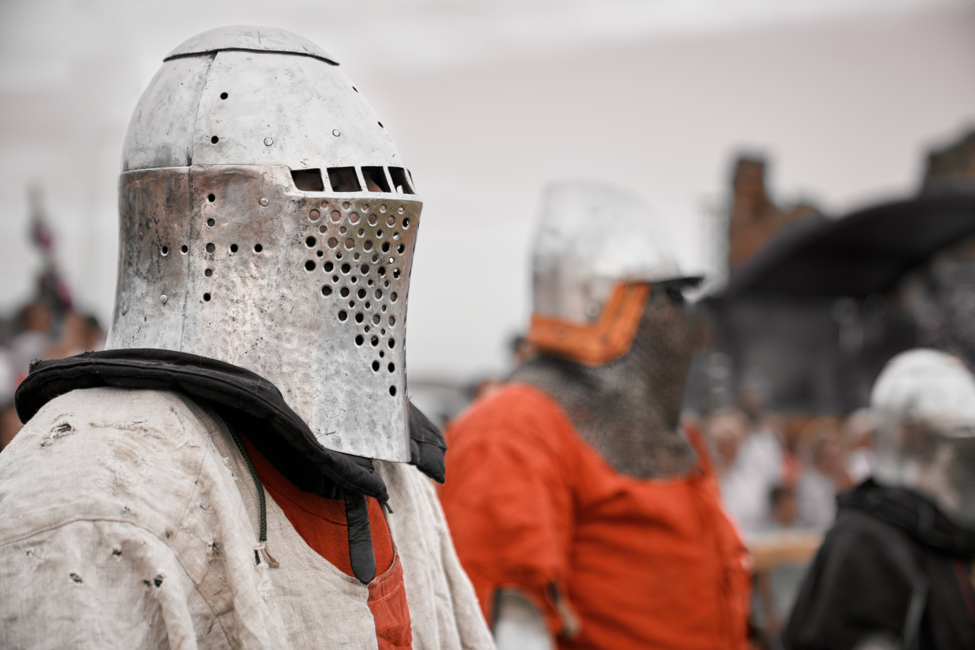 Two individuals dressed in medieval armor stand outdoors. The foreground figure wears a large metal helmet with a perforated visor and a padded, quilted tunic. In the background, another person wears similar armor, partially blurred.