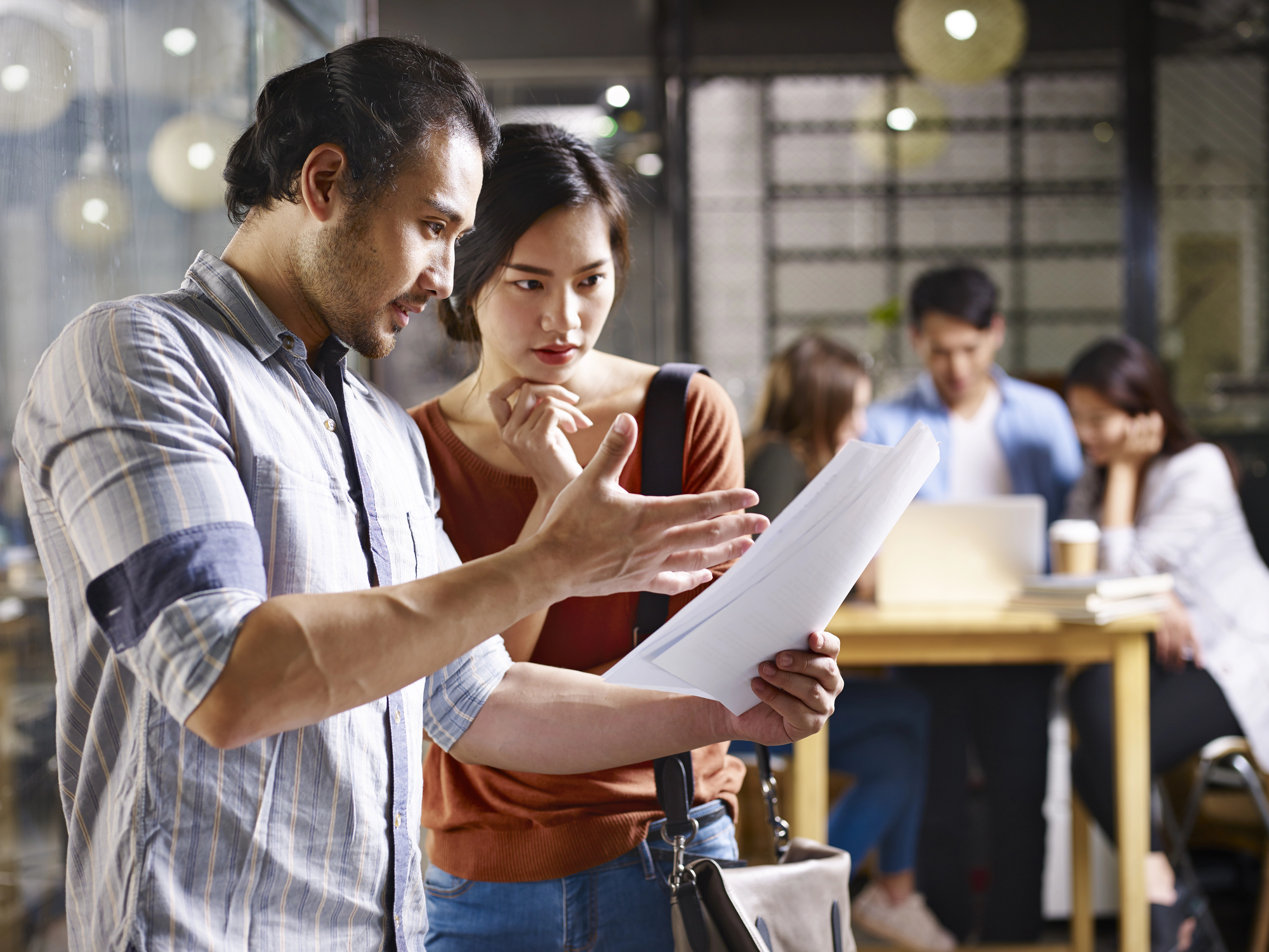 A man and woman stand in an office discussing documents. The man gestures while showing papers to the woman, who listens attentively. In the background, three other people work at a table with laptops and papers.