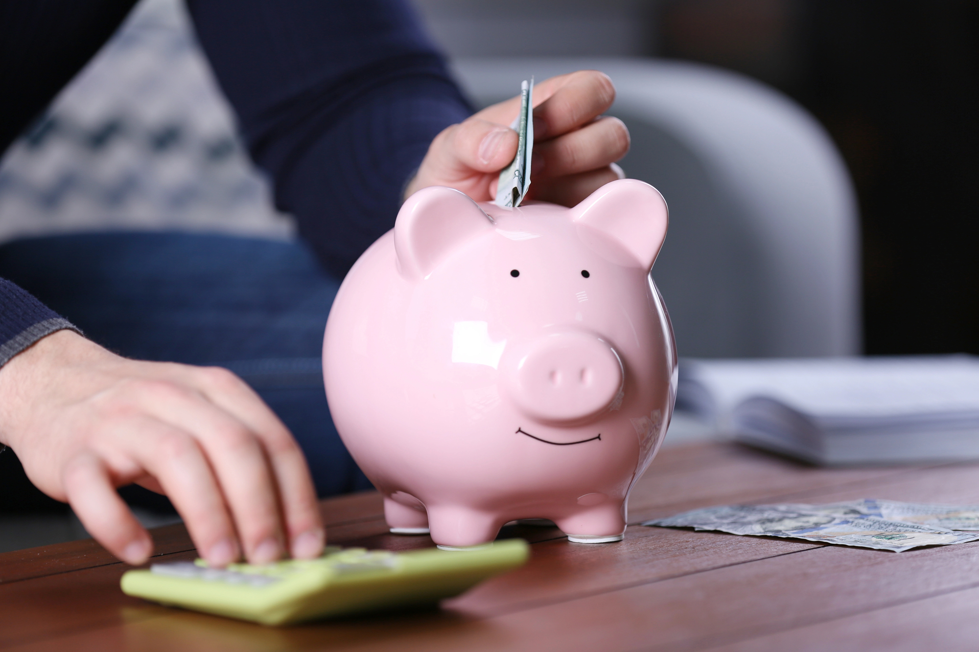 A person is placing money into a pink piggy bank on a wooden table. Their other hand is using a green calculator. Some scattered papers and money are visible in the background.