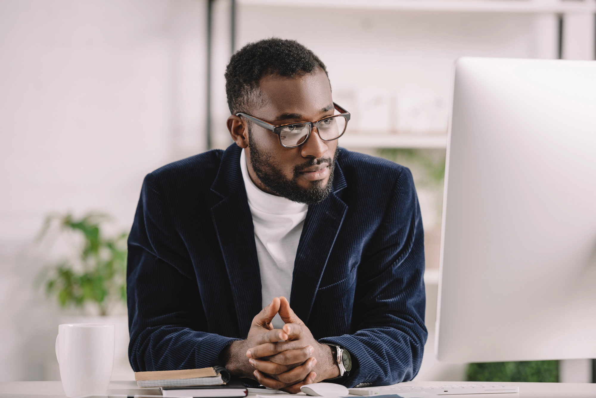 A man in glasses and a dark blazer leans forward, intently looking at a computer monitor. A notebook and a coffee mug sit on the desk. The background is softly lit, with shelves and a plant visible.