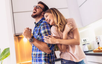 A man and woman stand back to back in a kitchen, both laughing joyfully. The man wears glasses and a plaid shirt, while the woman wears a pink top. The kitchen has white cabinets and a warm light from under the cabinets.