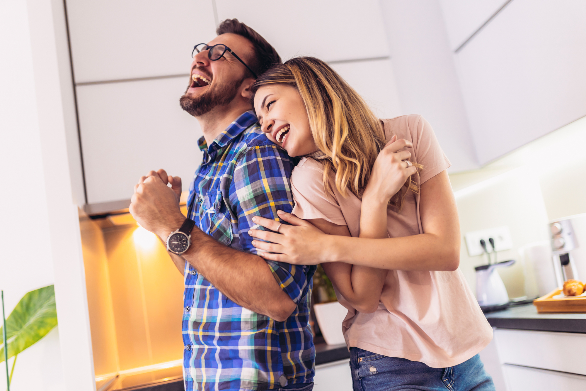 A man and woman stand back to back in a kitchen, both laughing joyfully. The man wears glasses and a plaid shirt, while the woman wears a pink top. The kitchen has white cabinets and a warm light from under the cabinets.