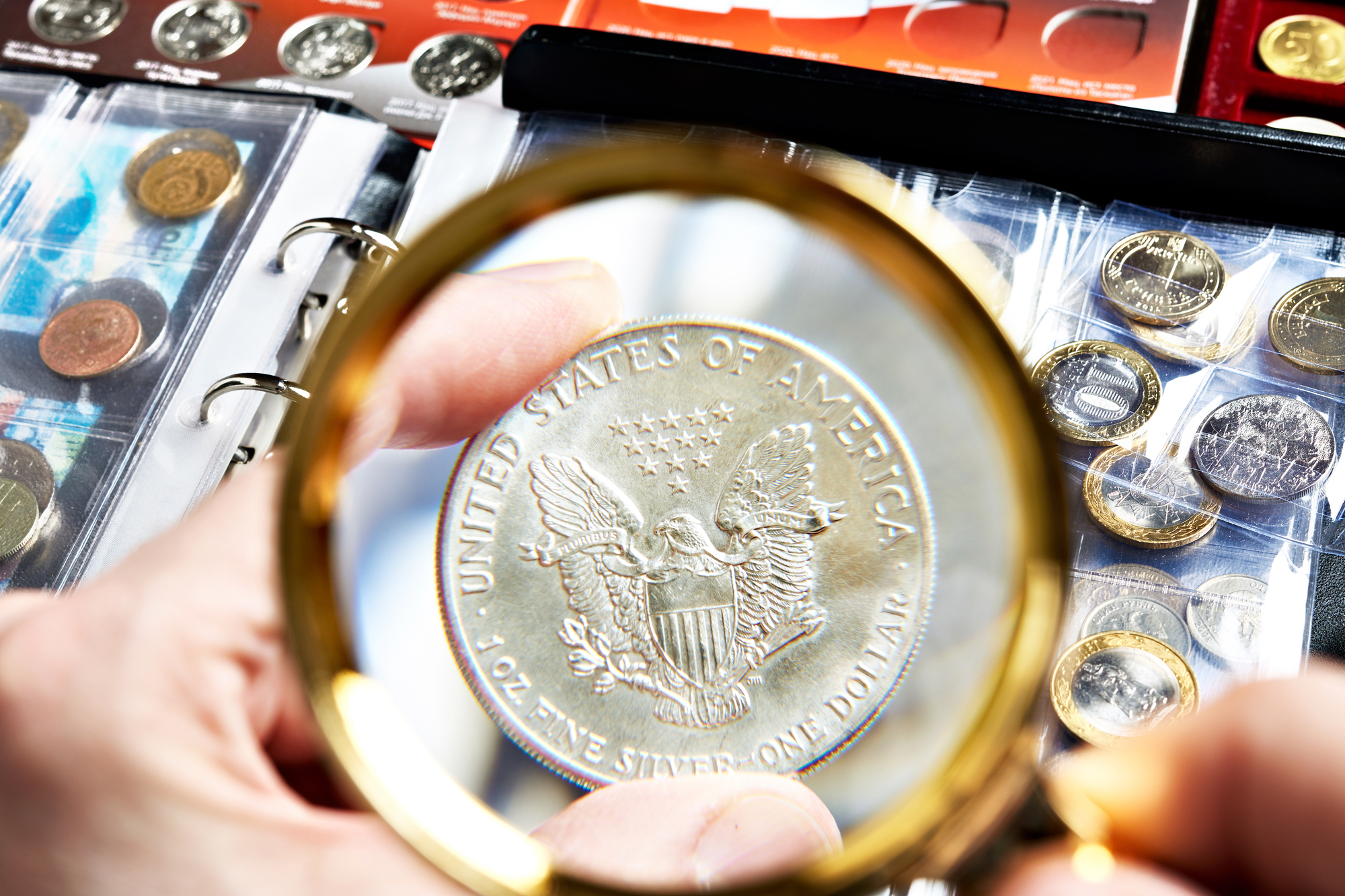 A person holds a magnifying glass over a U.S. silver coin featuring an eagle emblem. The scene includes an assortment of coins in an album in the background.