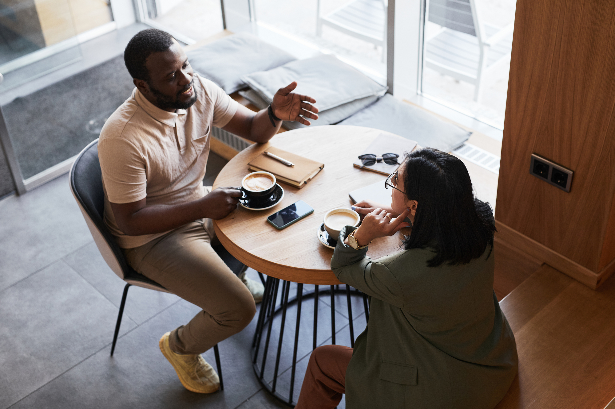 Two people sit at a cafe table, engaged in conversation. Each has a coffee and some personal items on the table, including a phone and a notebook. The atmosphere is casual and bright, with large windows in the background.