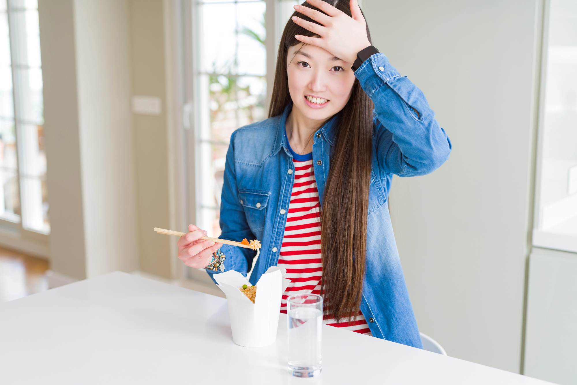 A person with long hair wearing a denim jacket and striped shirt sits at a table, holding chopsticks and eating from a takeout box. They have one hand on their forehead and a glass of water is nearby.