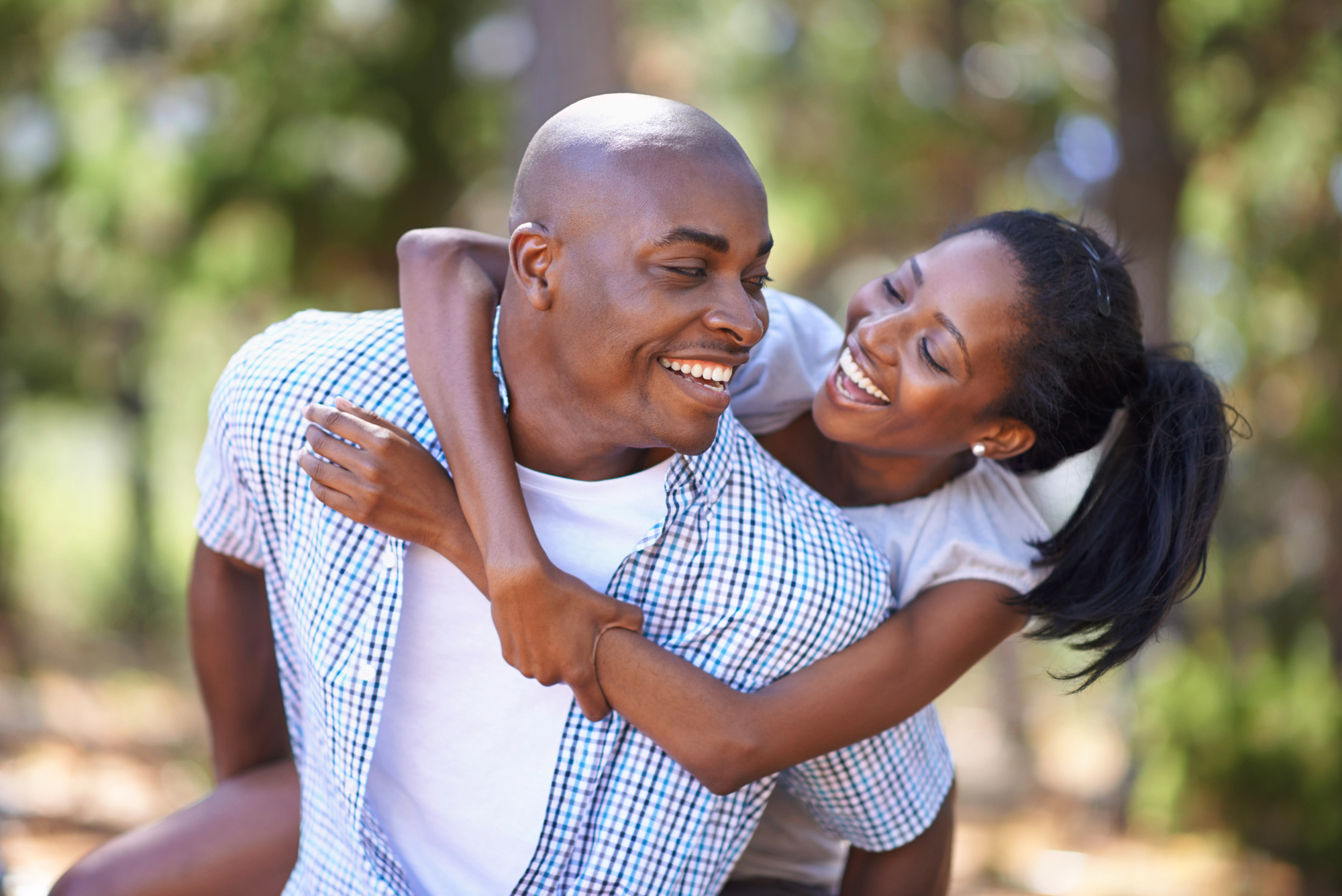 A person gives another person a piggyback ride outdoors. Both are smiling and appear to be enjoying a sunny day. The background is blurred with greenery, suggesting a park or natural setting.