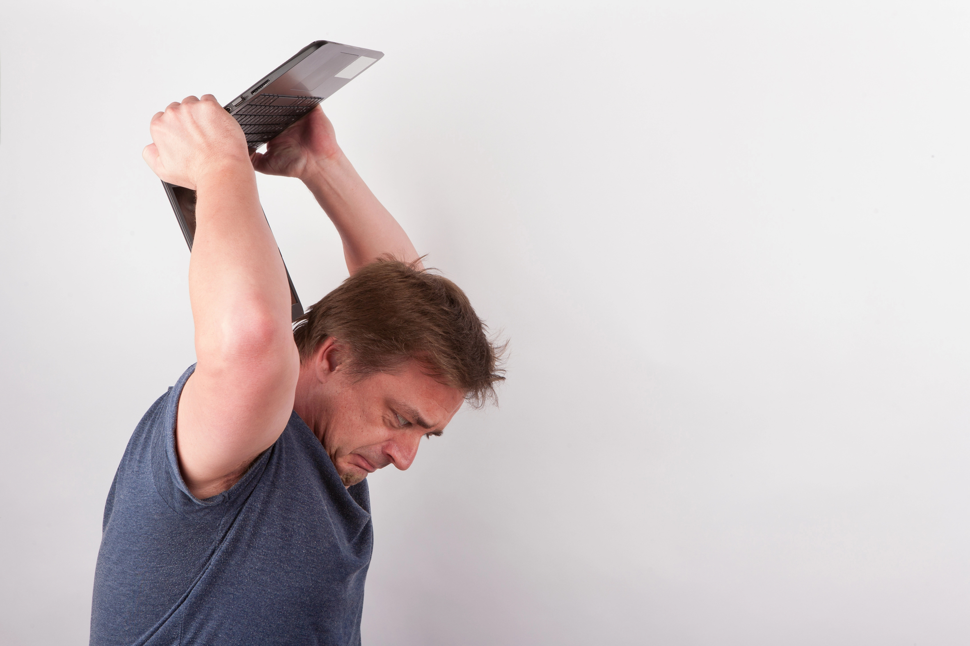 A man in a blue shirt holds a laptop overhead with both hands, appearing frustrated or angry, against a plain white background.