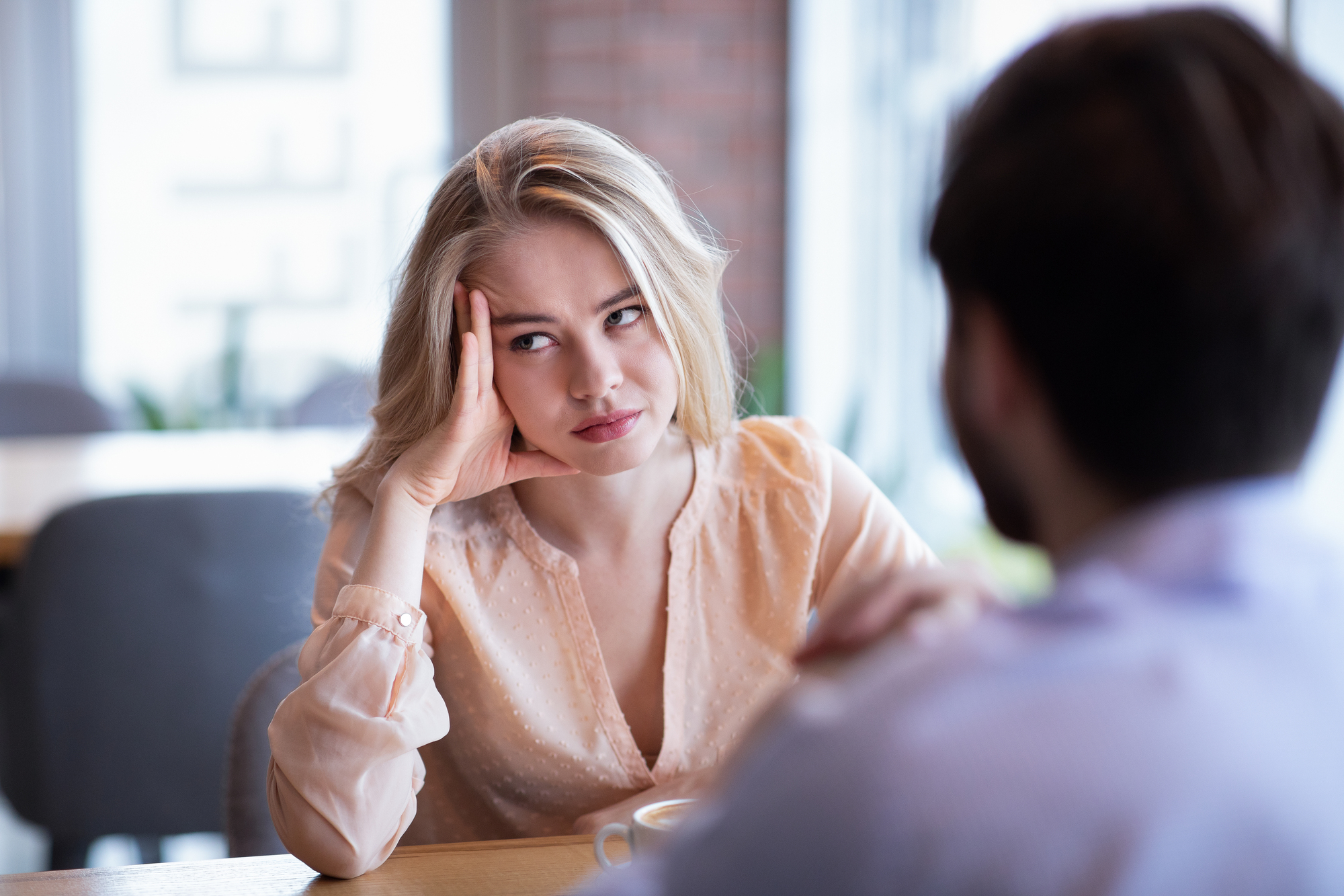 A woman with long blonde hair looks bored or annoyed, resting her head on her hand. She sits across from a man in a cafe. The background is slightly blurred, suggesting an indoor setting with natural light from large windows.