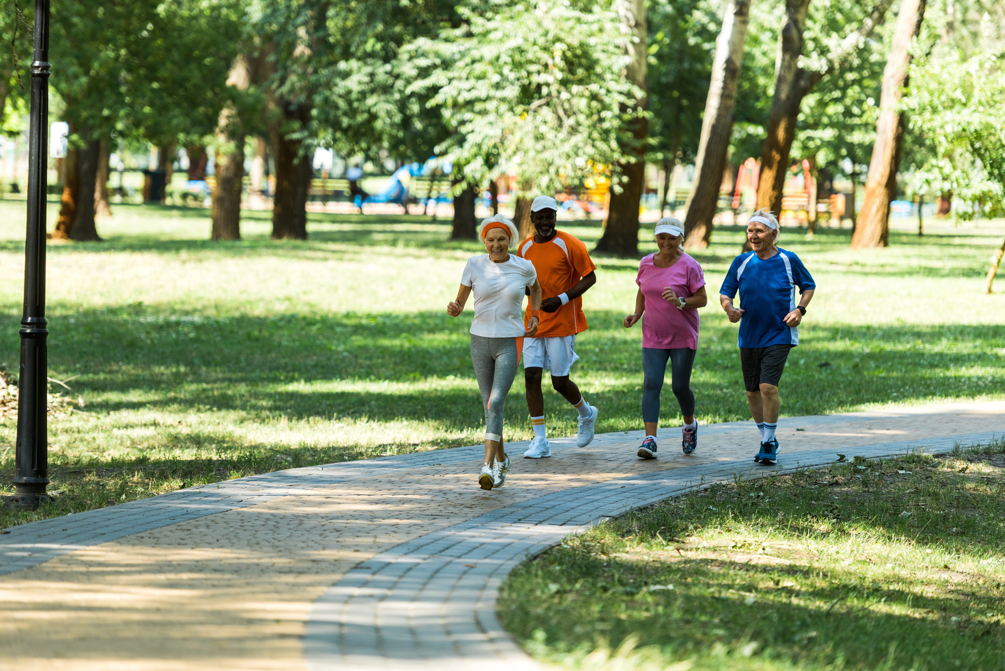 Four people are jogging together on a park path surrounded by lush green trees. They appear to be enjoying a sunny day, wearing comfortable athletic clothing and running shoes. The scene conveys a sense of fitness and camaraderie.