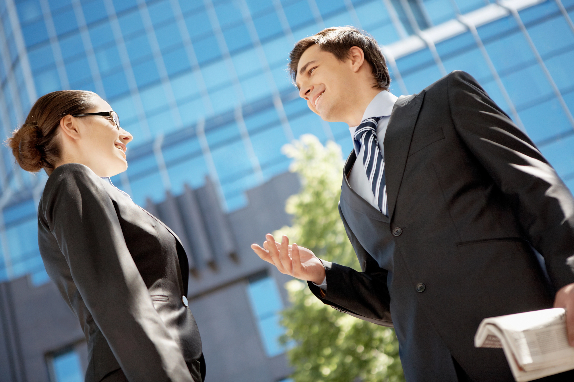 Two professionals in business attire converse outside an office building with a glass facade. The man holds a newspaper, and both appear to be smiling. A tree is visible in the background under a clear blue sky.