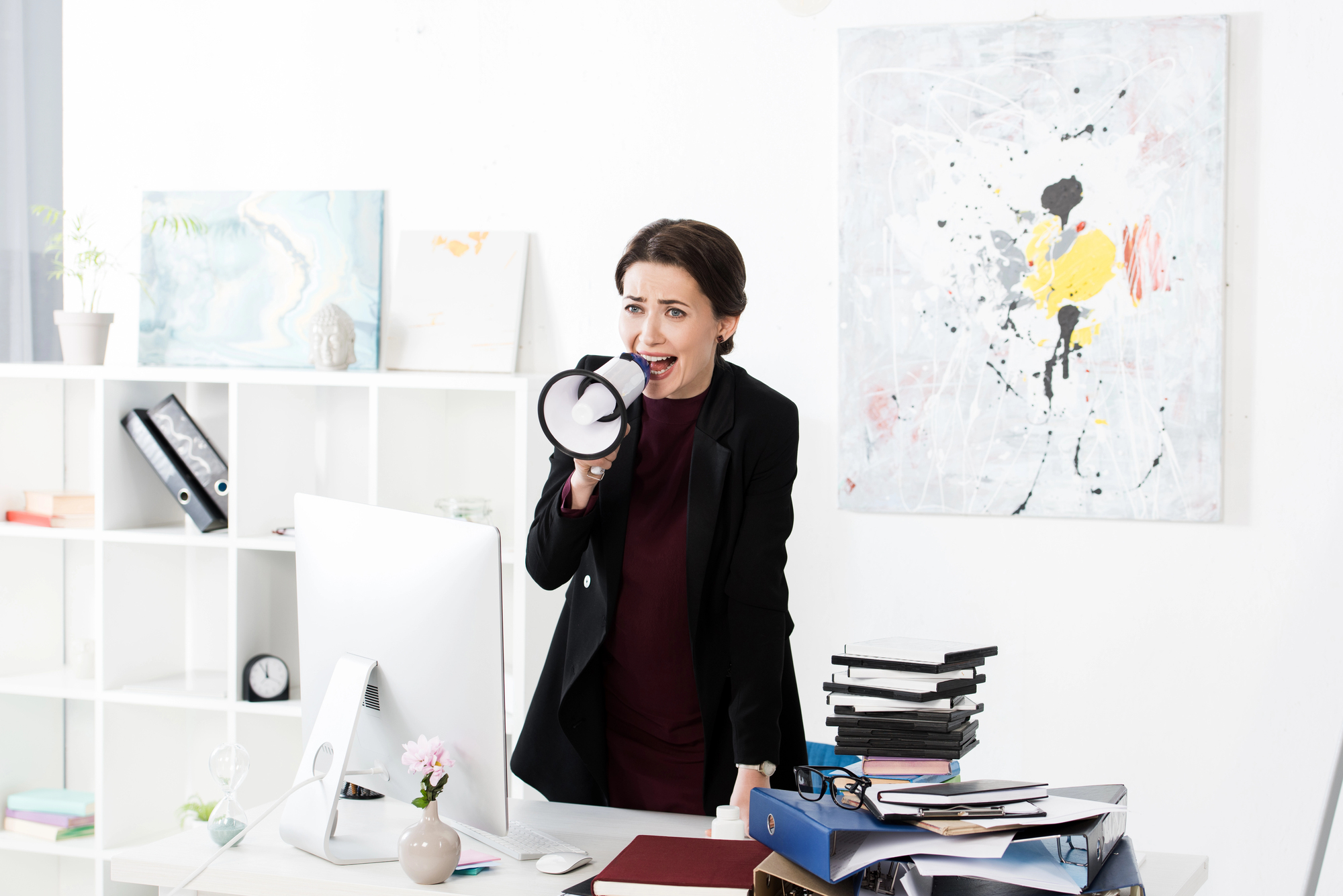 A woman in an office stands near a desk, speaking into a megaphone. She is dressed in a black blazer, surrounded by a computer, pile of files, and office decor. Abstract artwork hangs in the background.