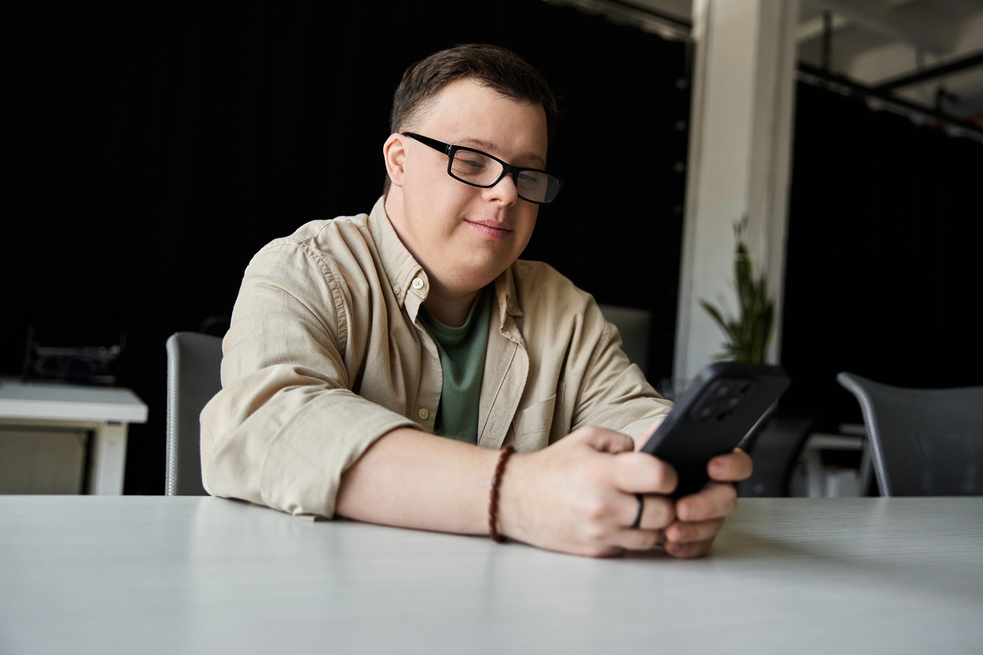 A person with short hair and glasses is seated at a table, smiling while looking at a smartphone. They are wearing a beige shirt over a green T-shirt, with a bracelet on one wrist. The background features a dark curtain and office chairs.