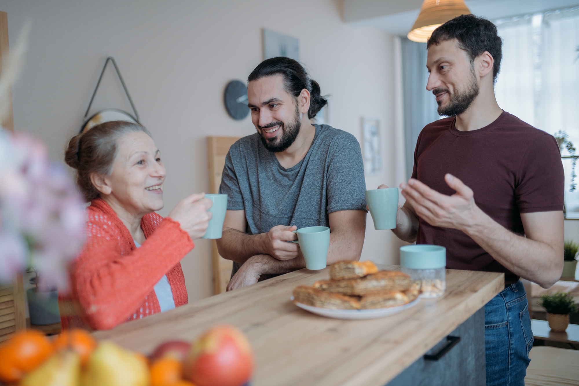 Three people enjoy a conversation while holding mugs in a cozy kitchen. A woman in an orange cardigan smiles while two men stand beside her, one gesturing with his hand. A plate of pastries and a bowl of fruit are on the table.