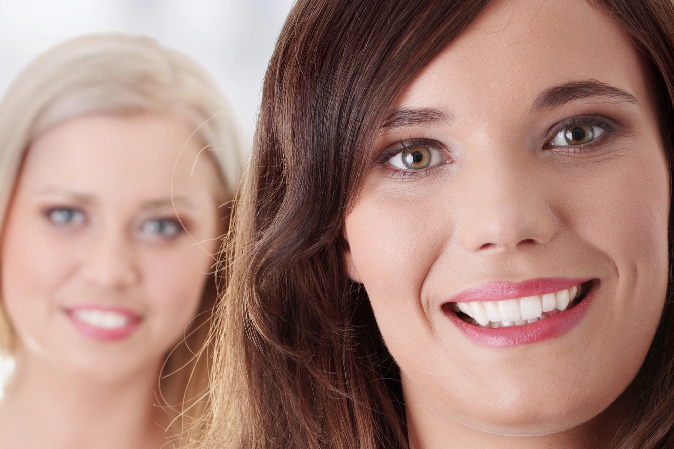 Two women smiling at the camera. The woman in the foreground has brown hair and is in focus, while the woman in the background has blonde hair and is slightly out of focus. The background is blurred, highlighting their expressions.