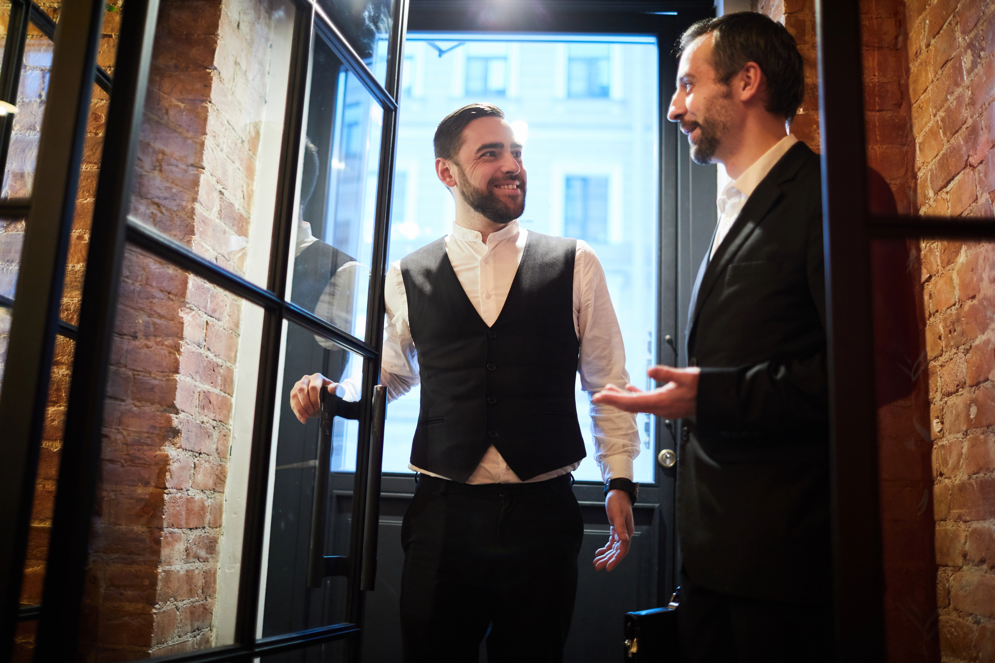 Two men in formal attire stand in a well-lit room with brick walls. One is holding a door, smiling at the other, who gestures with one hand. They appear to be engaged in conversation. The setting has a modern, industrial feel.