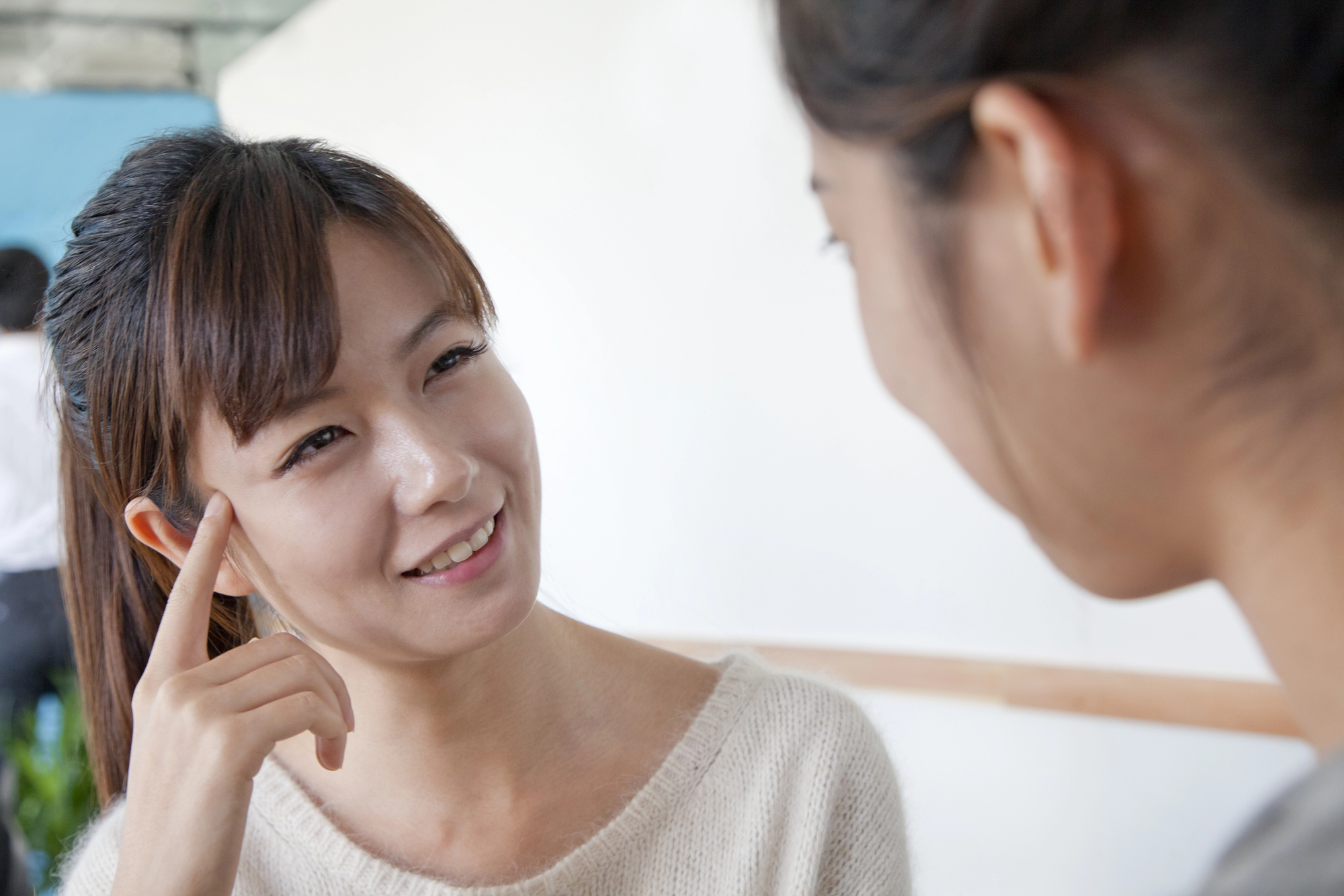 Two women are engaged in a friendly conversation. One woman is smiling and touching her temple, wearing a white sweater, while the other is blurred in the foreground. The setting appears to be casual and indoors.