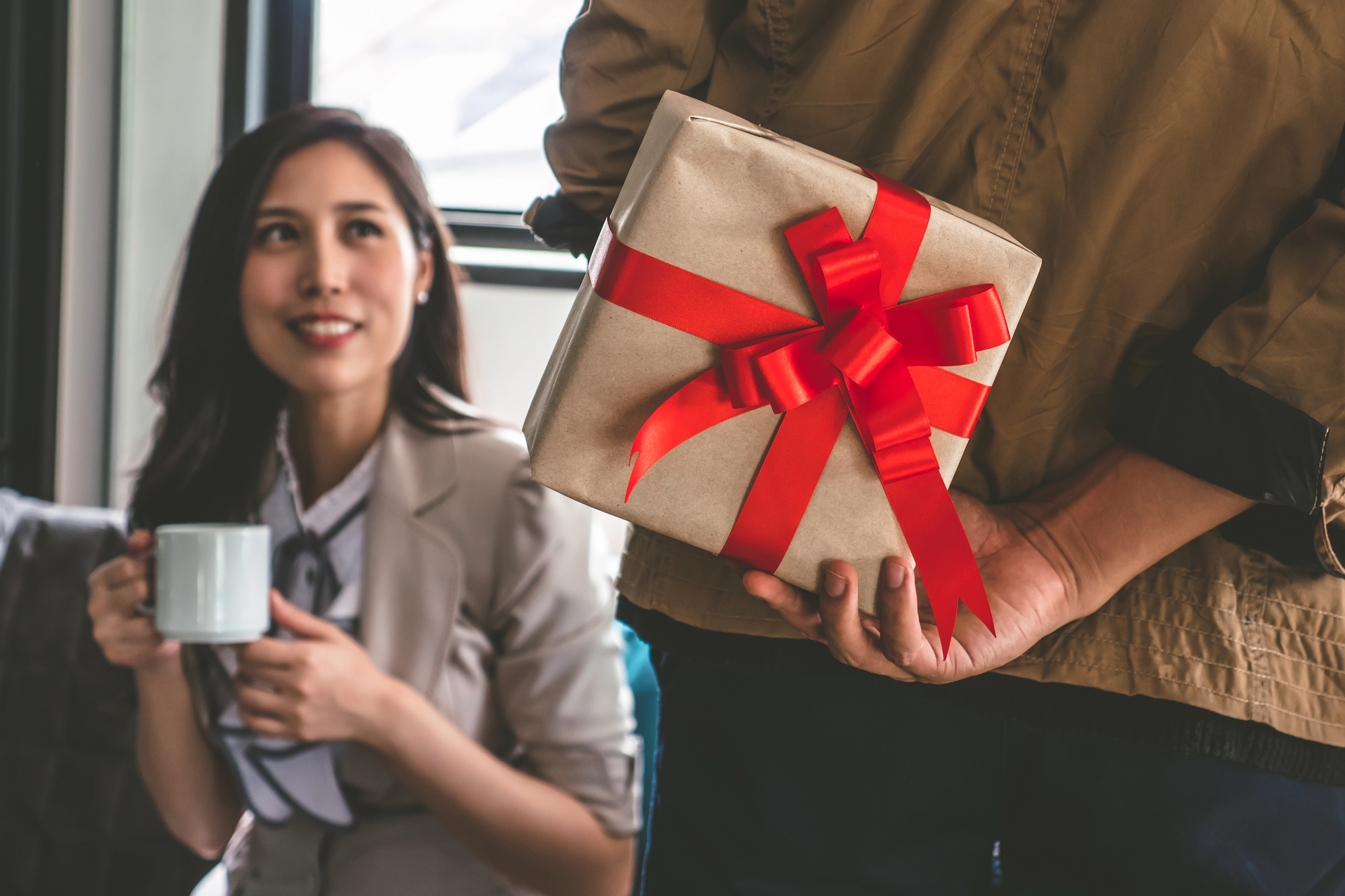 A person holds a gift wrapped in brown paper with a red ribbon behind their back, while facing a smiling person holding a cup. The scene suggests a surprise gift scenario indoors.