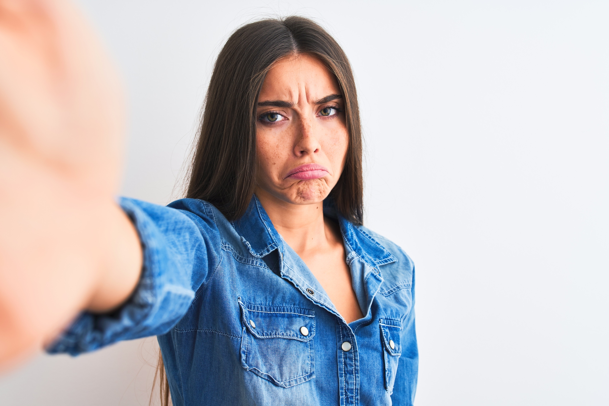 A woman with long brown hair wearing a denim shirt is making a pouty face. Her arm is extended towards the camera as if taking a selfie. The background is plain, off-white.