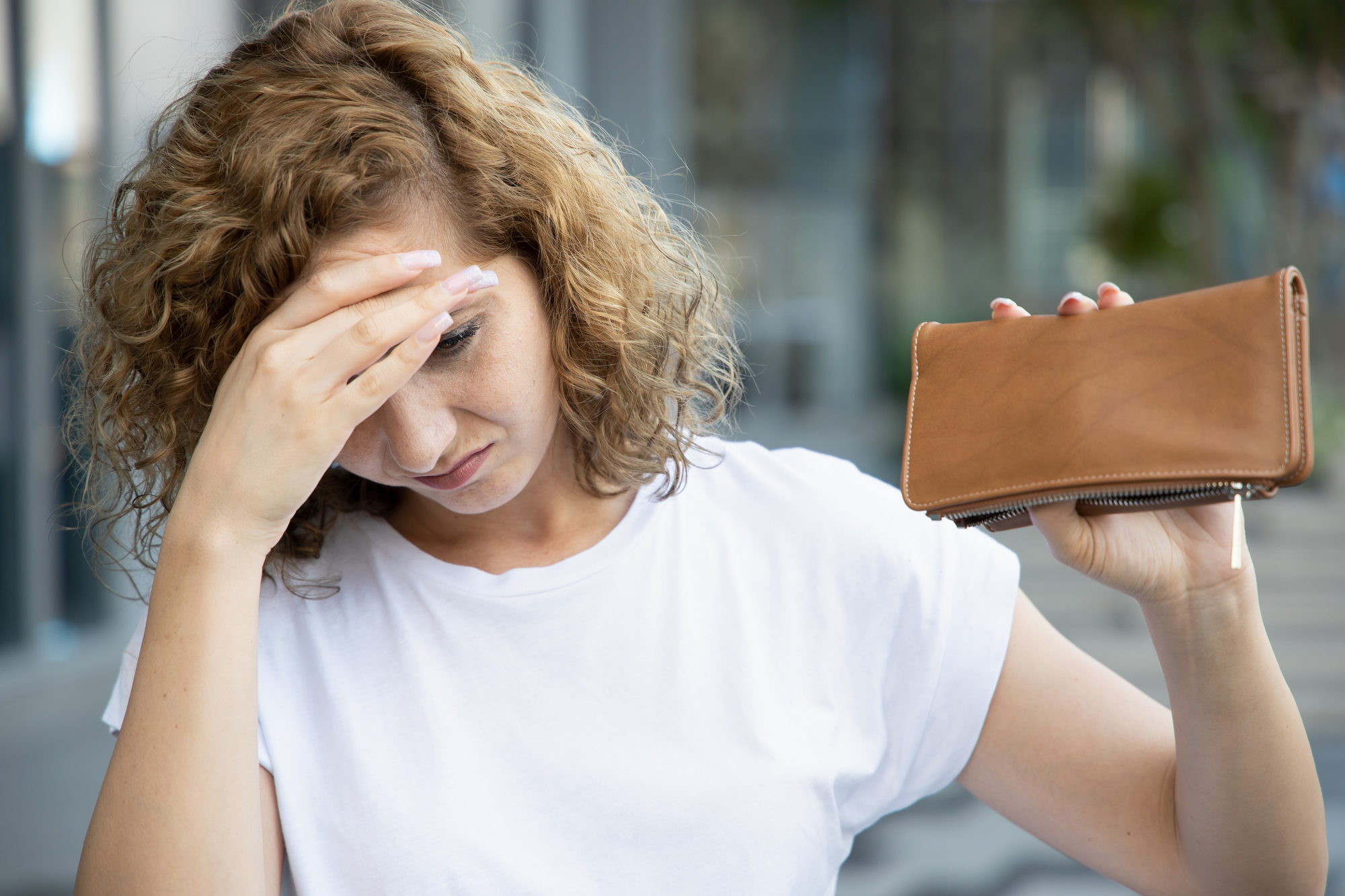 A woman with curly hair looks distressed, holding her forehead with one hand and an empty brown wallet in the other. She is wearing a white shirt and standing outdoors.