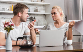 A man and a woman are sitting at a kitchen table with a laptop and documents. They appear to be having a serious discussion. A coffee pot and cup are on the table. The kitchen is modern with open shelves.