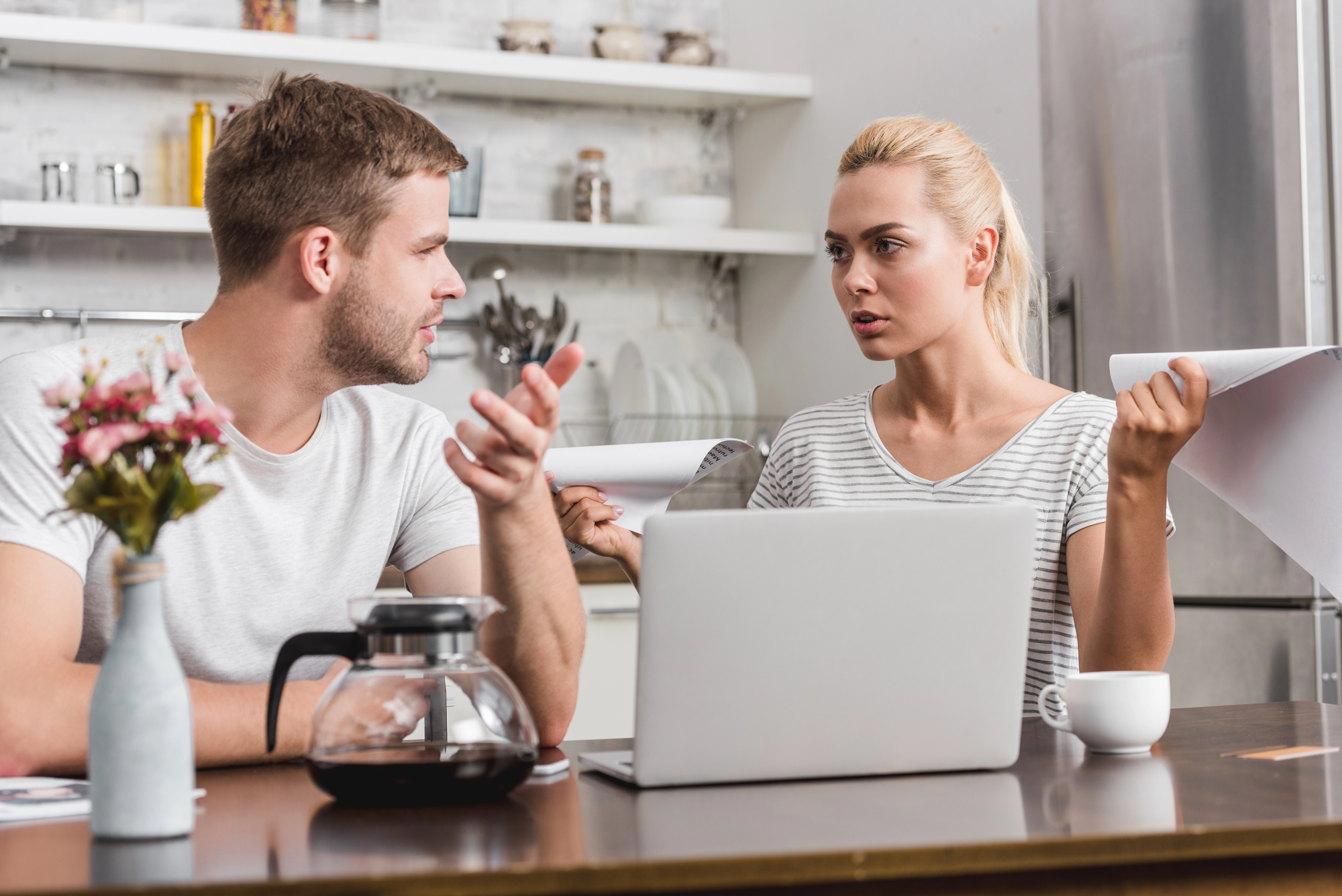 A man and a woman are sitting at a kitchen table with a laptop and documents. They appear to be having a serious discussion. A coffee pot and cup are on the table. The kitchen is modern with open shelves.