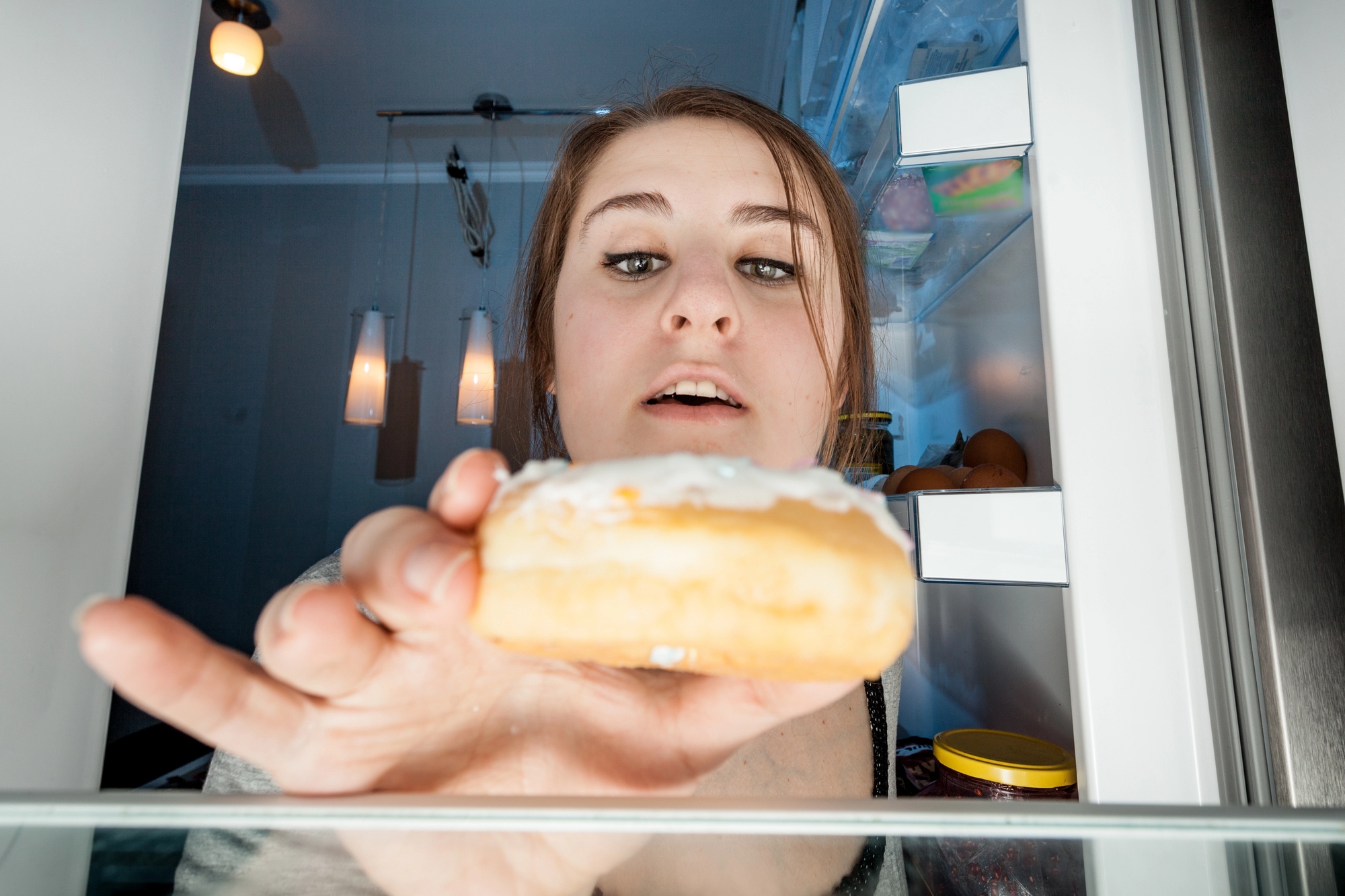 A woman reaches into an open refrigerator, looking at a frosted donut in her hand. The fridge is visible, containing some jars and produce. The kitchen background includes hanging lights.