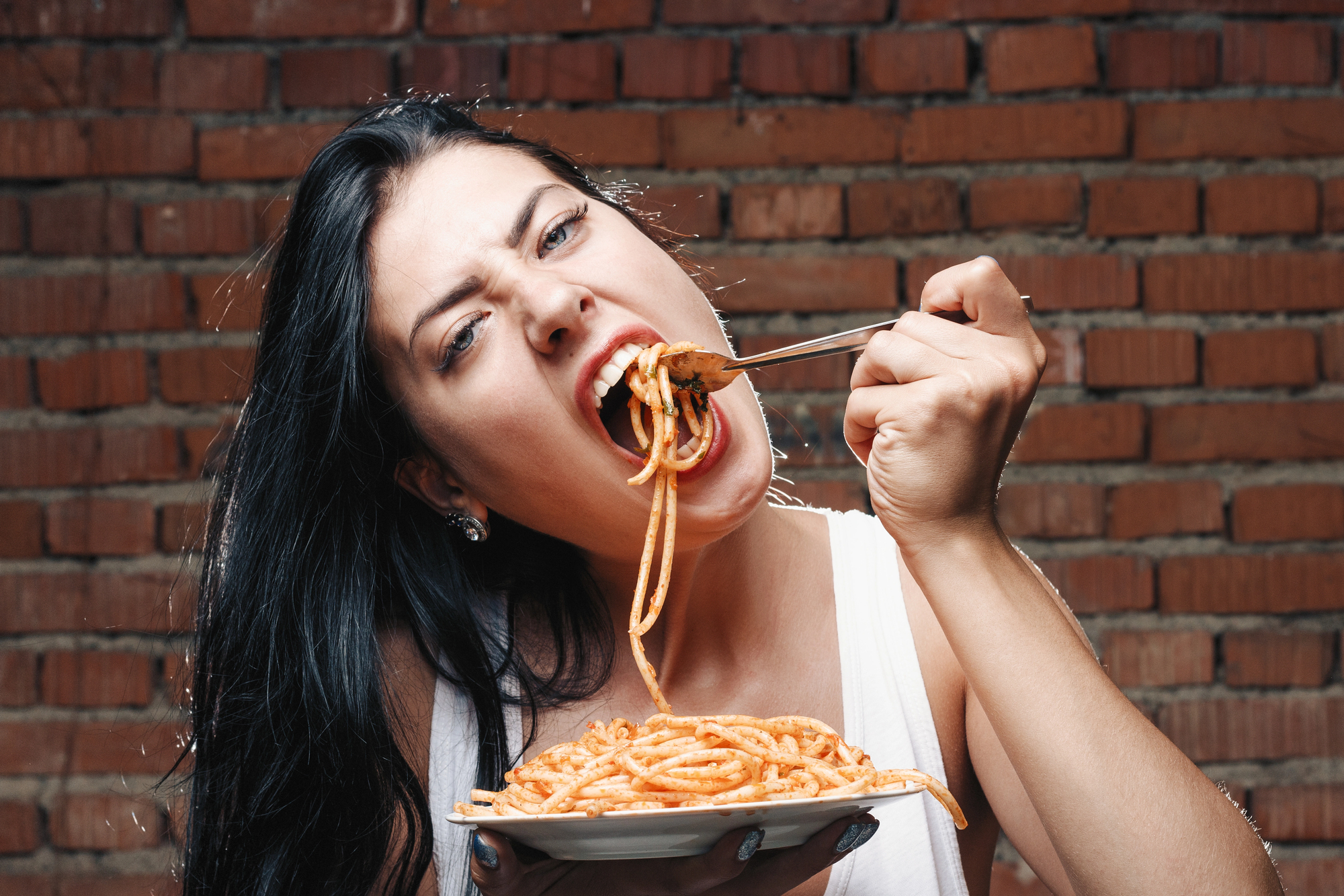 A woman with long dark hair enthusiastically eating spaghetti from a fork against a brick wall background. She is wearing a white tank top and holding a plate of pasta. Her expression is playful and energetic.