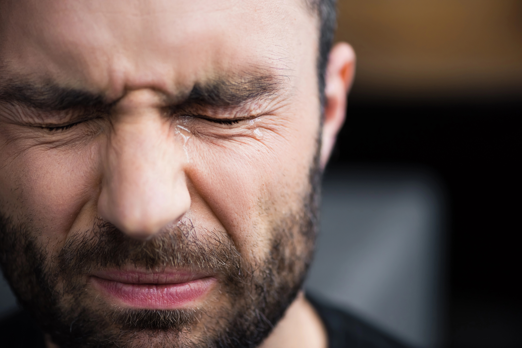 Close-up of a bearded man with dark hair, wearing a black shirt, and eyes tightly shut. His forehead is slightly wrinkled, and mouth pursed, suggesting discomfort or deep concentration. The background is softly blurred.