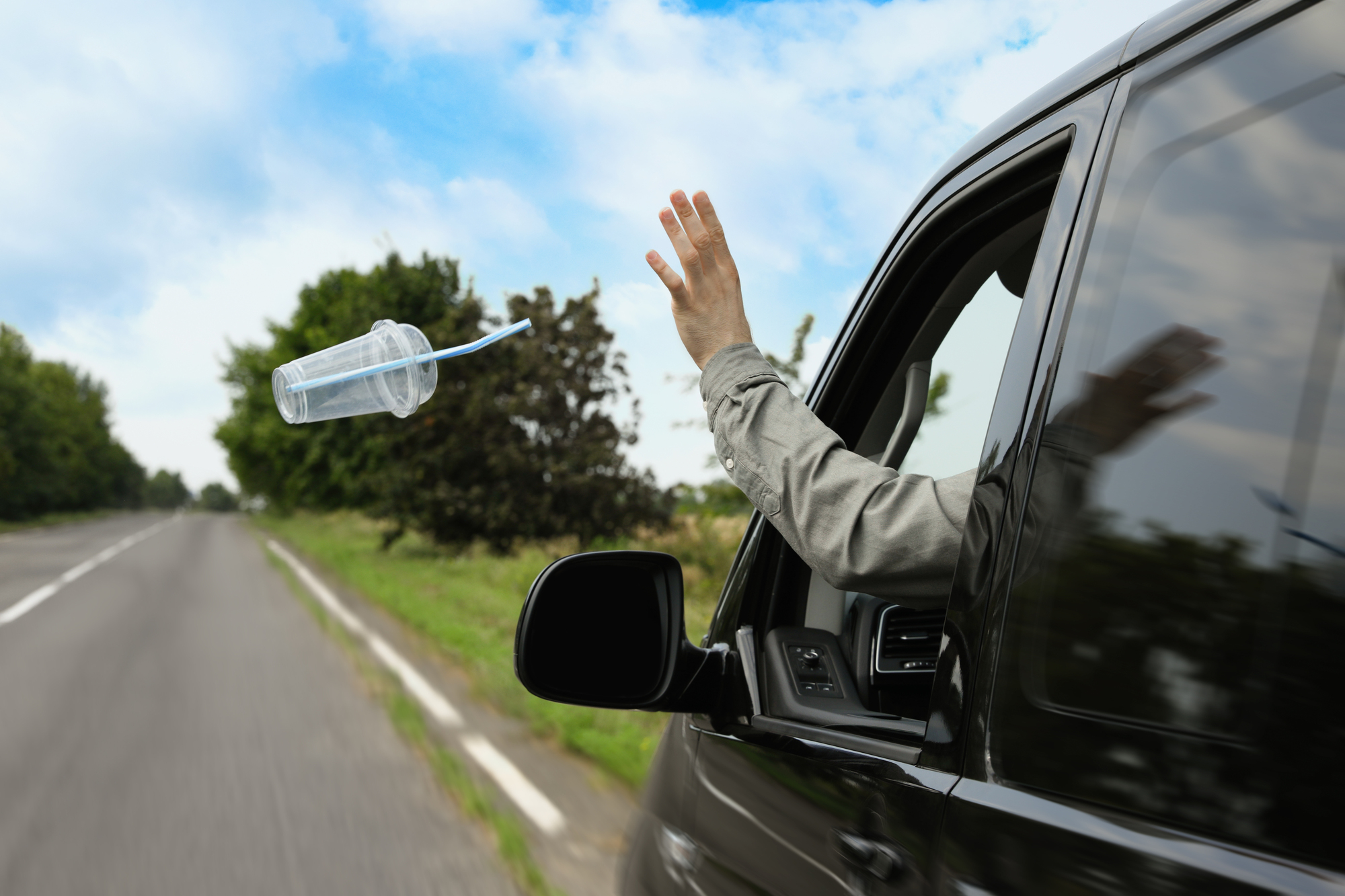 A hand is shown tossing a plastic cup with a straw out of a car window on a rural road. Trees and a blue sky are in the background, highlighting an act of littering.