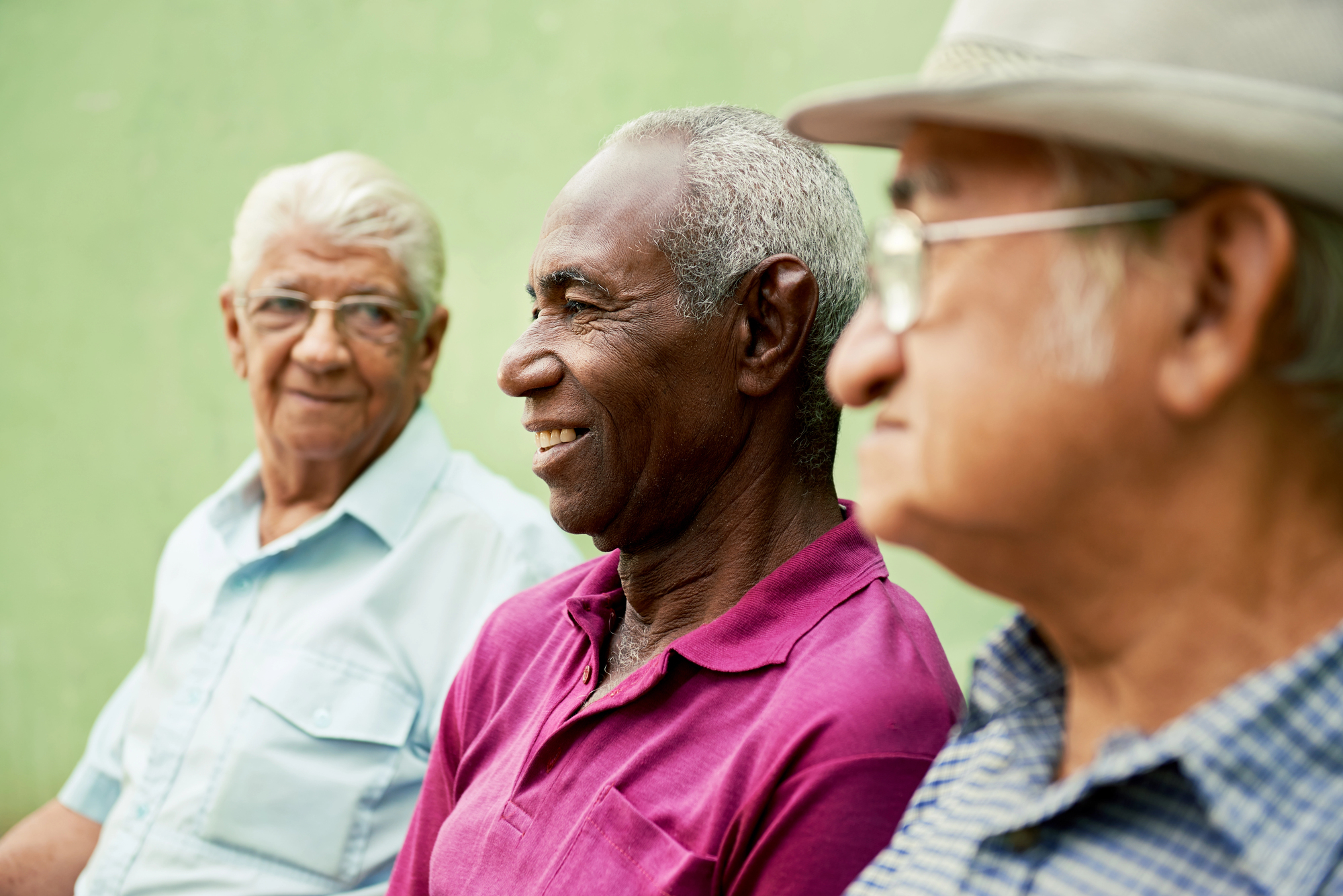 Three elderly men sitting outdoors, smiling and talking. The man in the middle, wearing a burgundy shirt, is facing forward. The other two men, in light shirts, are looking at him. They appear to be enjoying a friendly conversation against a green background.