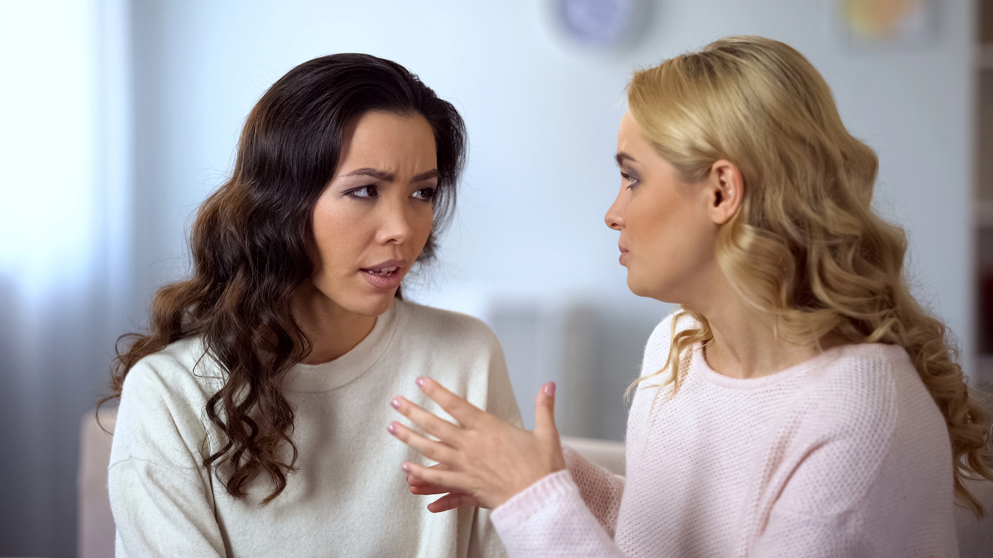 Two women are sitting indoors having a serious conversation. The woman on the left looks concerned, while the woman on the right gestures with her hands, appearing to explain something. Both are wearing light-colored sweaters.