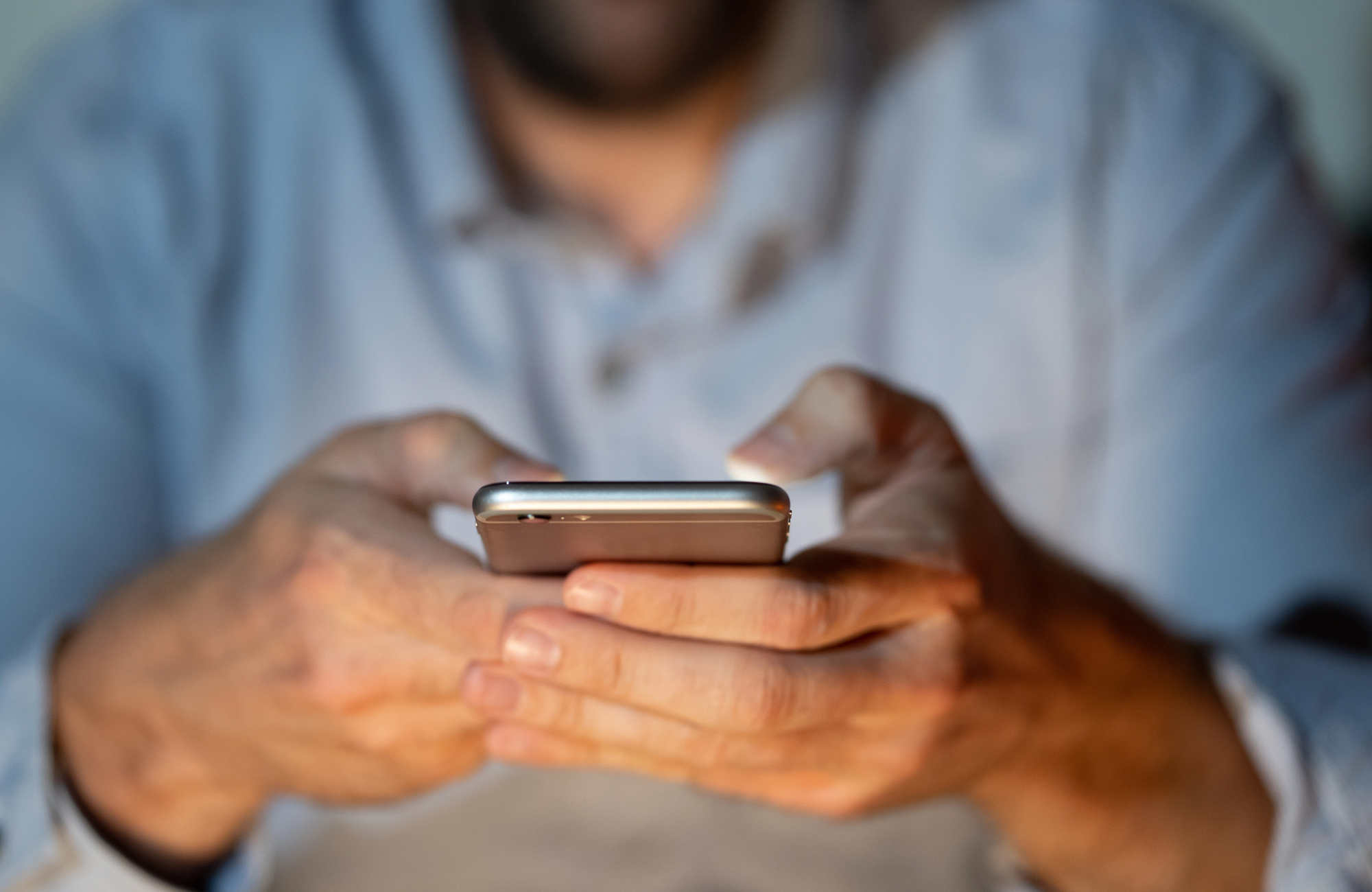 A person holding a smartphone with both hands, focusing on the screen. The background is blurred, and the person is wearing a light-colored shirt.