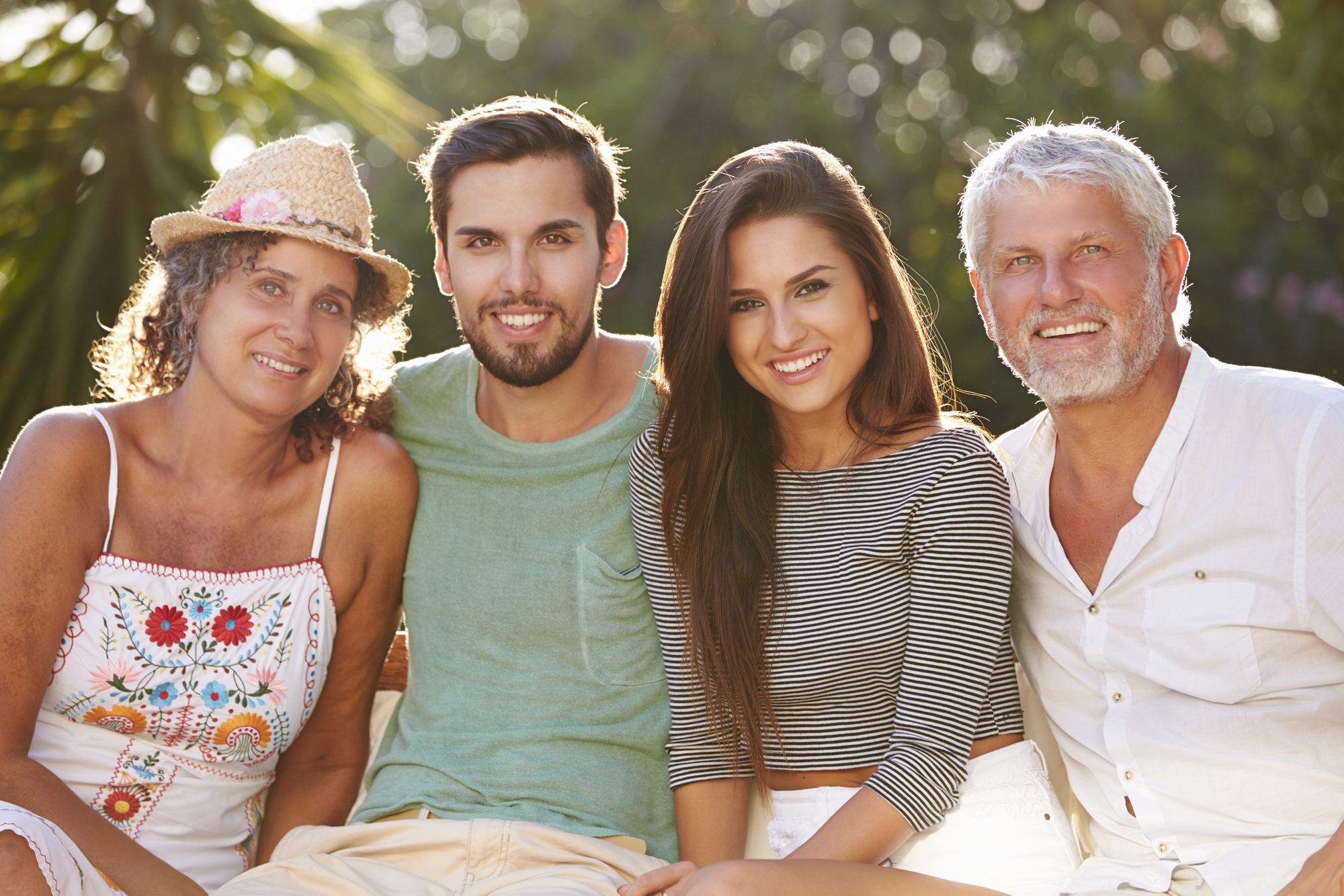 A group of four people sitting closely together outdoors. They are smiling and appear happy, surrounded by greenery. The older woman is wearing a hat, and the younger man is in a green shirt. The younger woman wears a striped top, and the older man is in white.