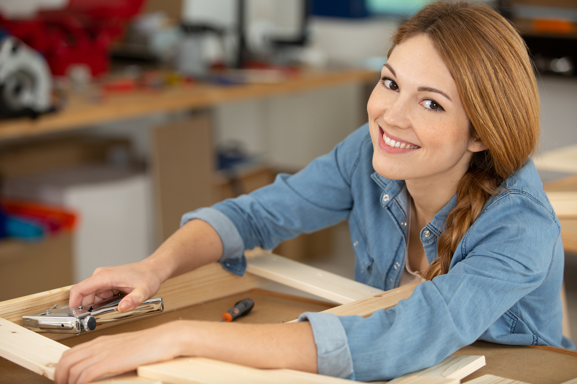 A smiling woman in a workshop is assembling a wooden frame. She wears a denim shirt and has red hair pulled into a braid. A stapler and screwdriver are on the table. The background shows various tools and workbenches.