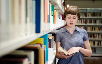 A person with short hair, wearing a blue collared shirt, stands in a library, reading a book. They are surrounded by tall shelves filled with books.